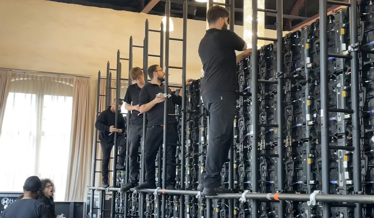 Four technicians standing on scaffolding working on large LED screens at an event setup.