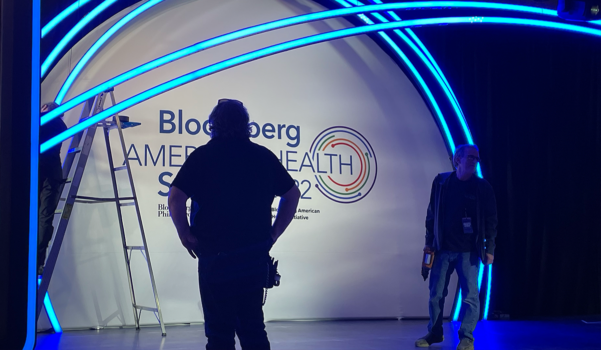 Silhouetted figures setting up at Bloomberg American Health Summit stage with ladder and futuristic blue light arch.