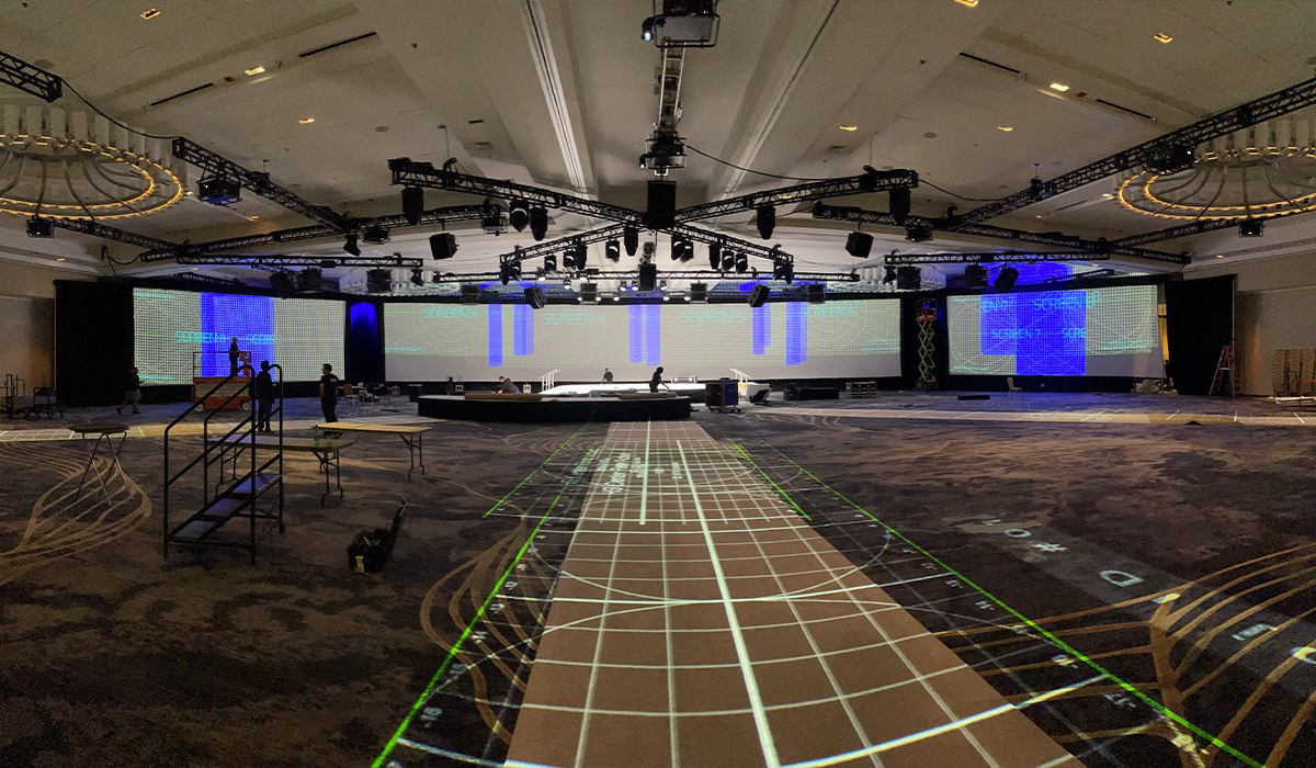 Large conference room with a stage and multiple LED screens, with crew members setting up lighting and equipment.
