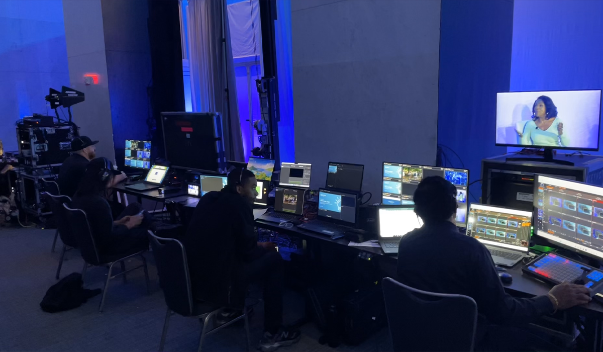 A control room with multiple computer monitors and operators working, with a large screen displaying a woman speaking on the right.