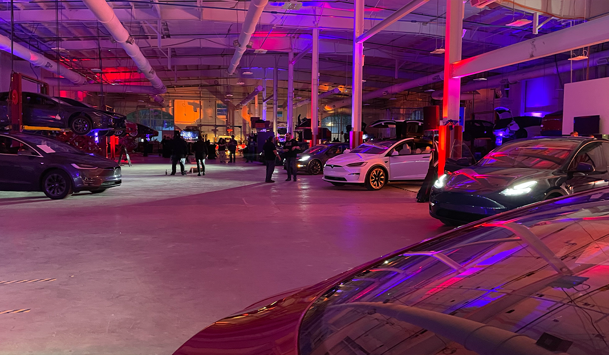 Indoor car showroom with multiple Tesla vehicles on display and people walking around, illuminated by colorful lighting effects.