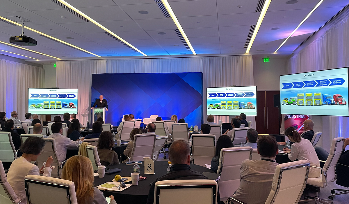 A conference room with a speaker presenting on stage, two large screens displaying a business vision, and an audience seated at round tables.