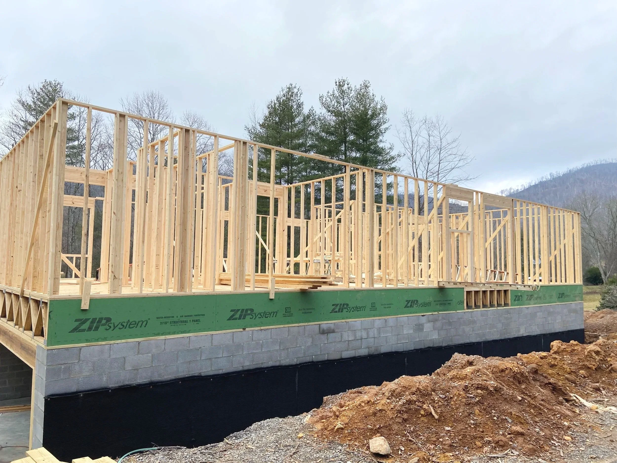 Wooden framing of a house under construction on concrete foundation with a view of trees and mountains in the background.