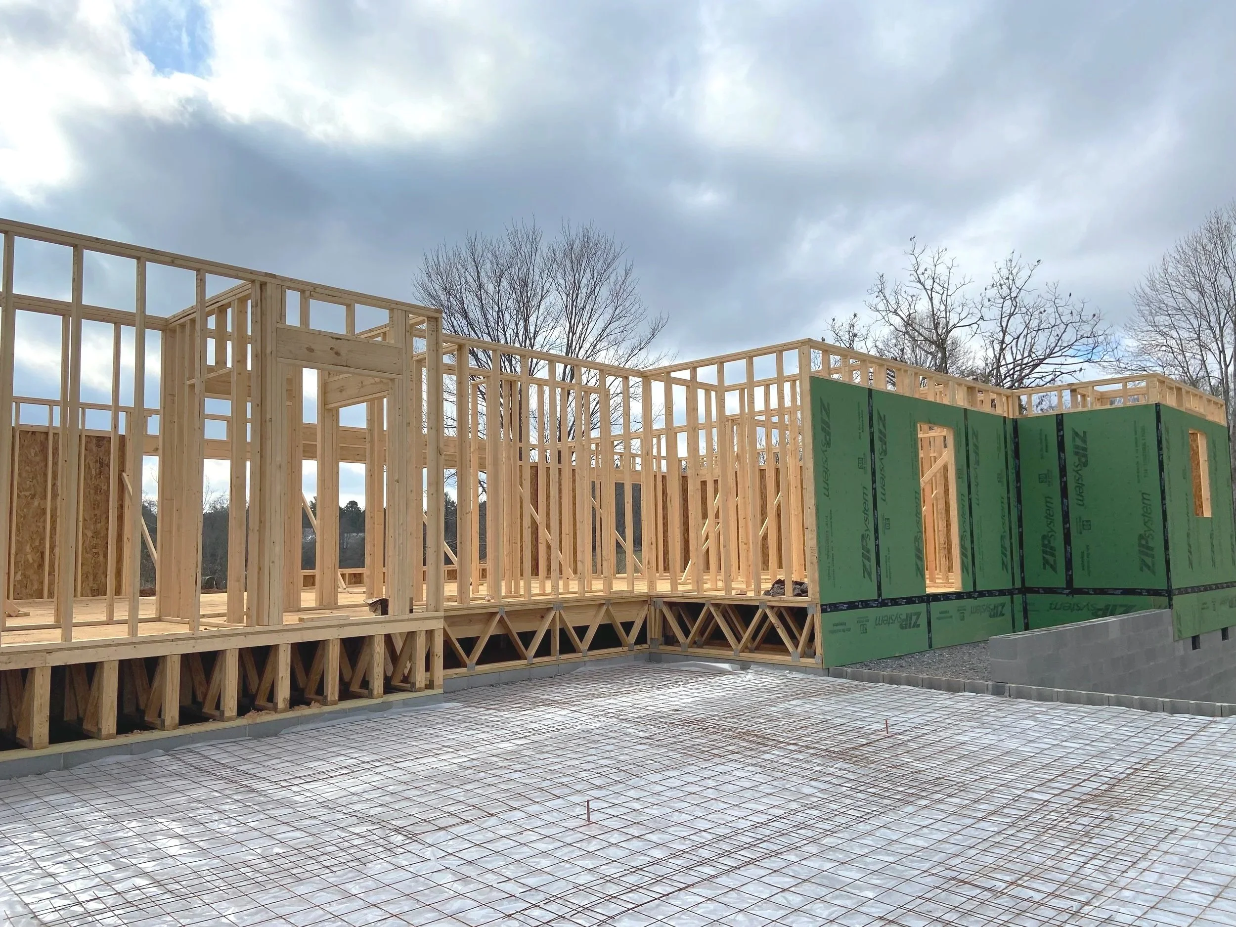Construction site with wooden framing for building walls, partially covered with green sheathing, and a concrete foundation with rebar grid.