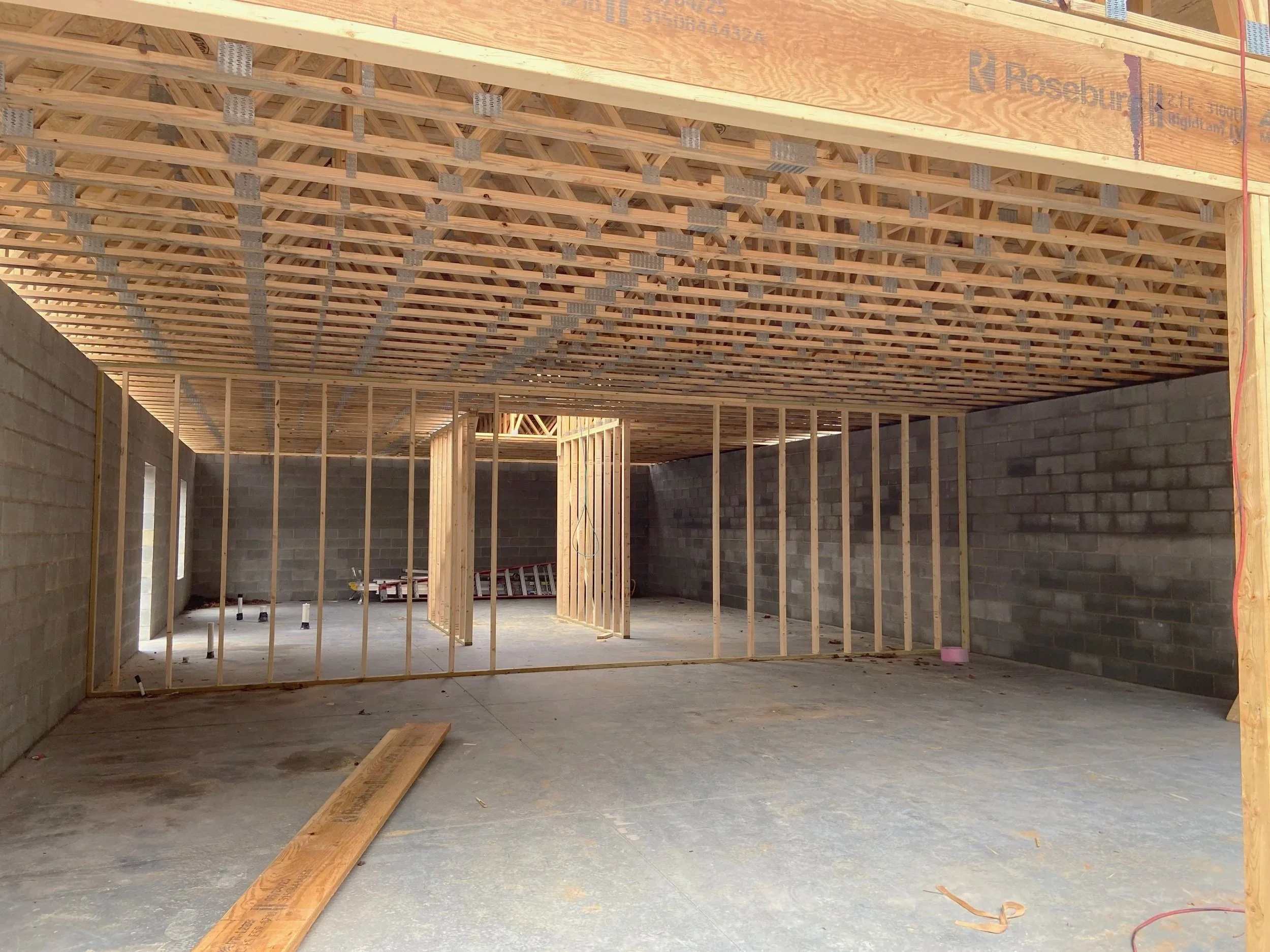 Construction site showing the framed interior of a building with wooden studs and roof trusses, unfinished walls, and a concrete floor.