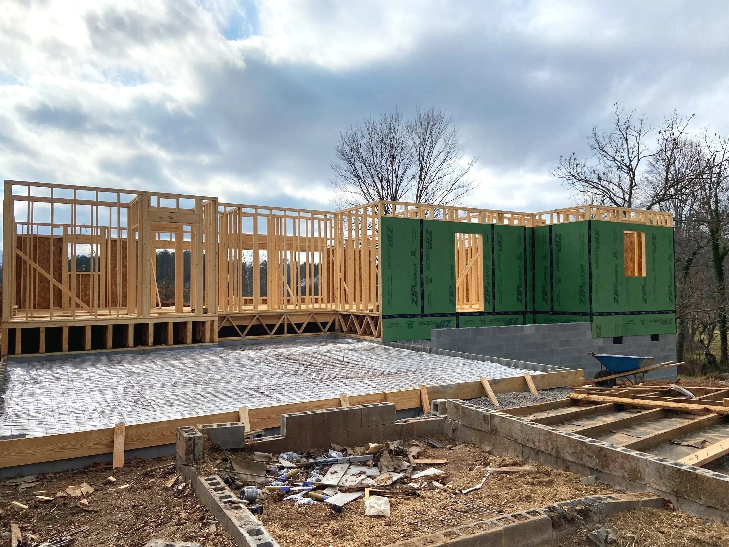 Construction site with wooden framing for a house, partially built with green sheathing on some walls, and a concrete foundation, with overcast sky and leafless trees in the background.