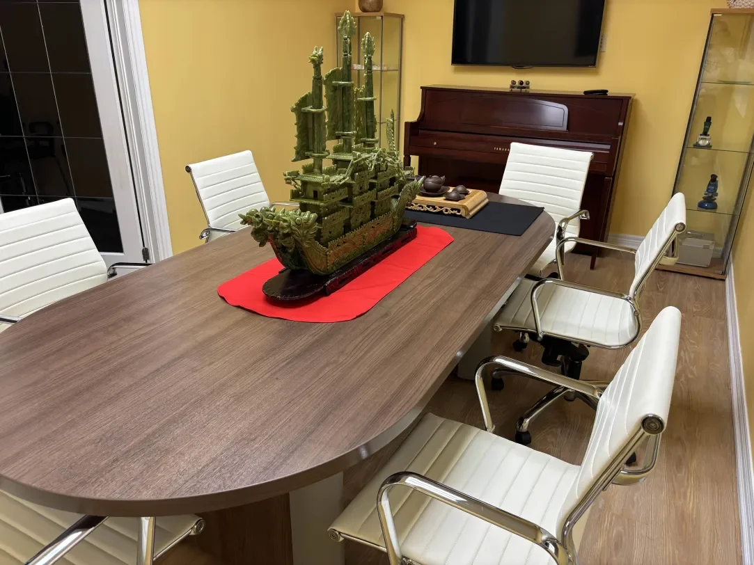A conference room with a wooden table, white ergonomic chairs, a decorative ship centerpiece, a red cloth underneath it, a western-style upright piano, a flat-screen TV on the yellow wall, and glass cabinets with decorative items.