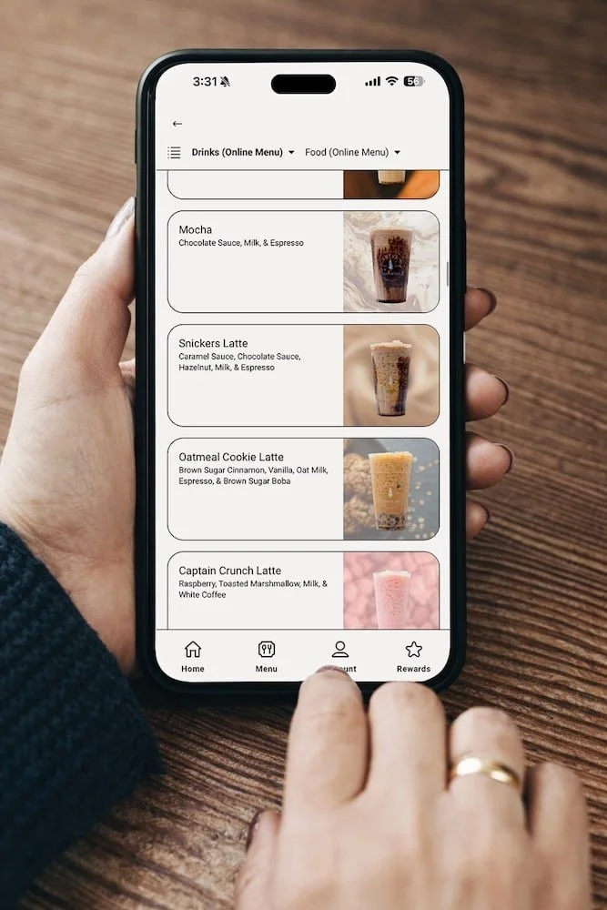 Mobile phone displaying a digital menu for coffee drinks, including mocha, Snickers latte, oatmeal cookie latte, and captain crunch latte, held over a wooden table.