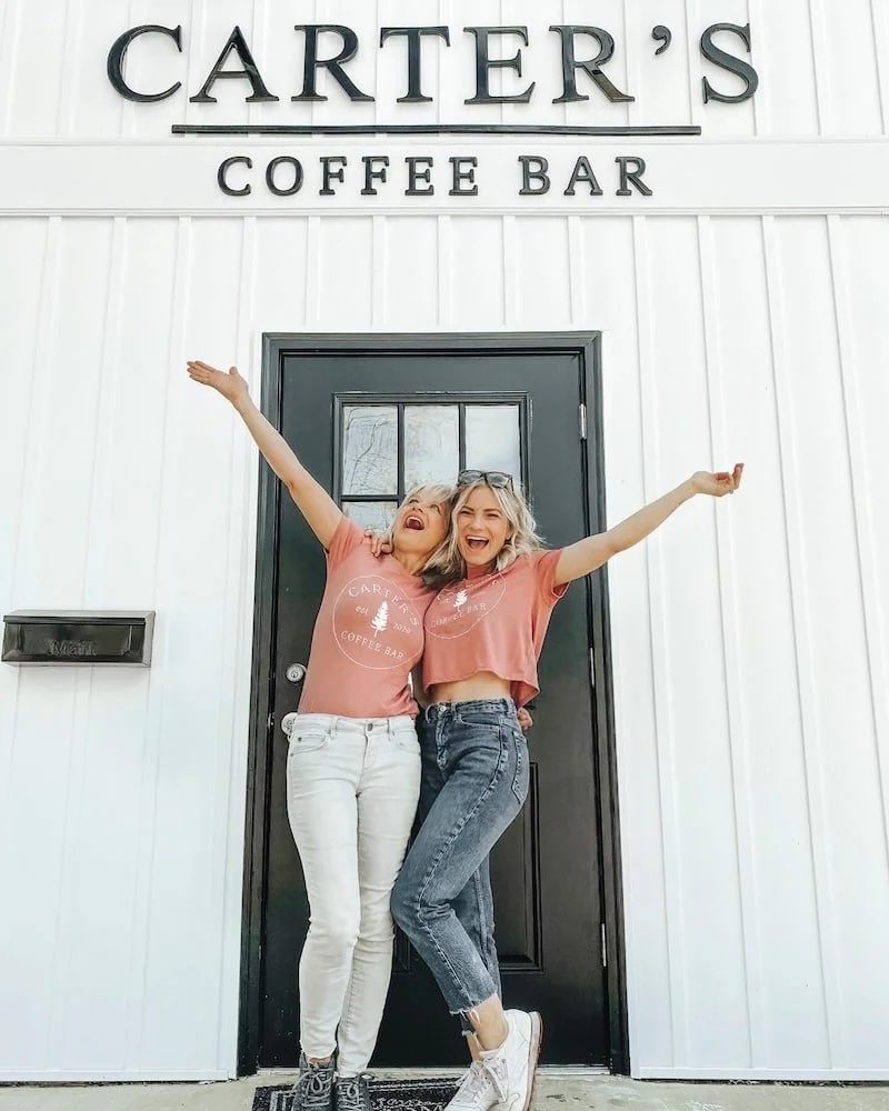 Two women standing in front of Carter's Coffee Bar, smiling and celebrating with arms raised.