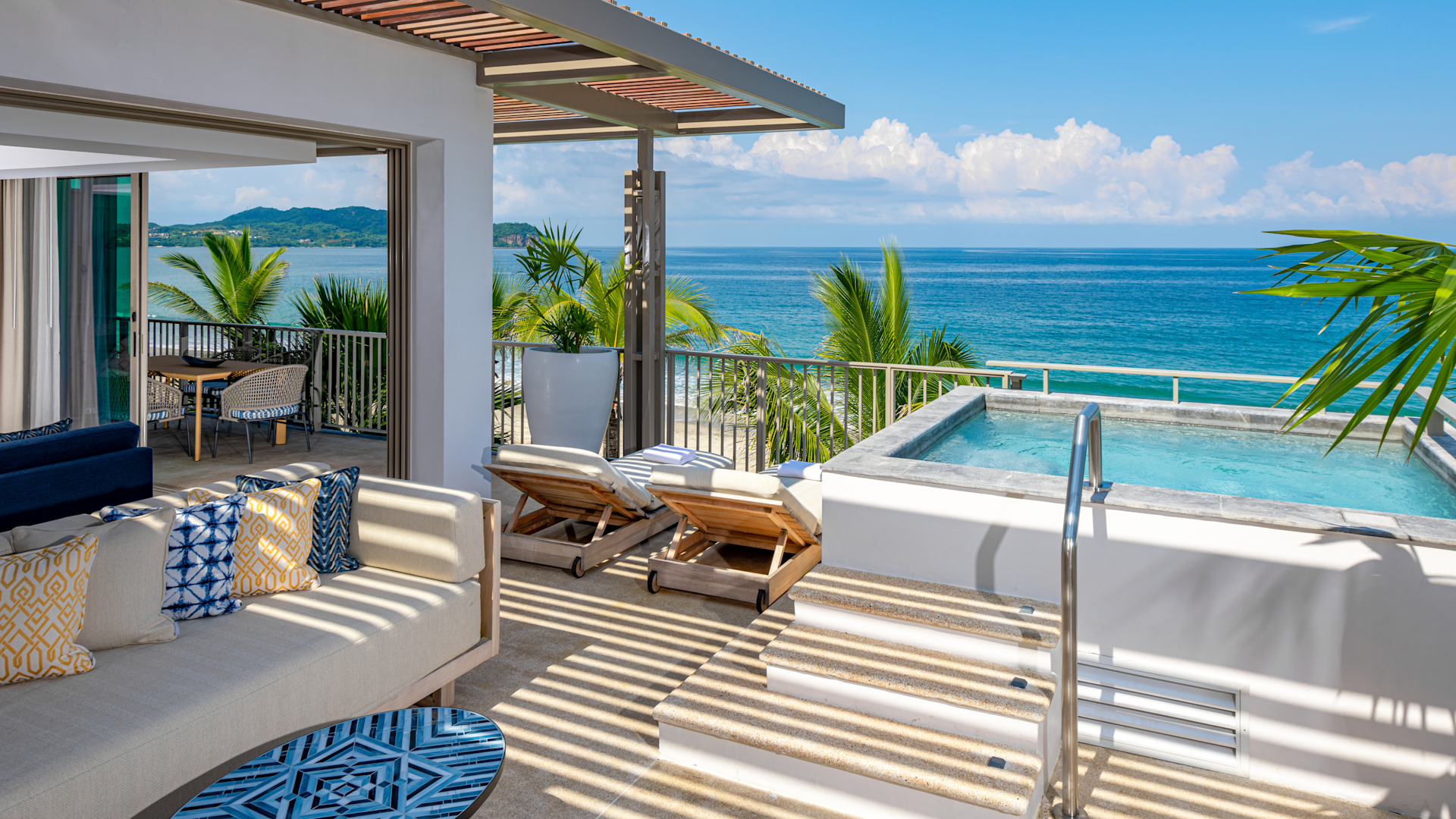 Luxury oceanfront balcony with outdoor seating, sun loungers, a hot tub, and tropical plants, overlooking a beach and the ocean with clear skies.