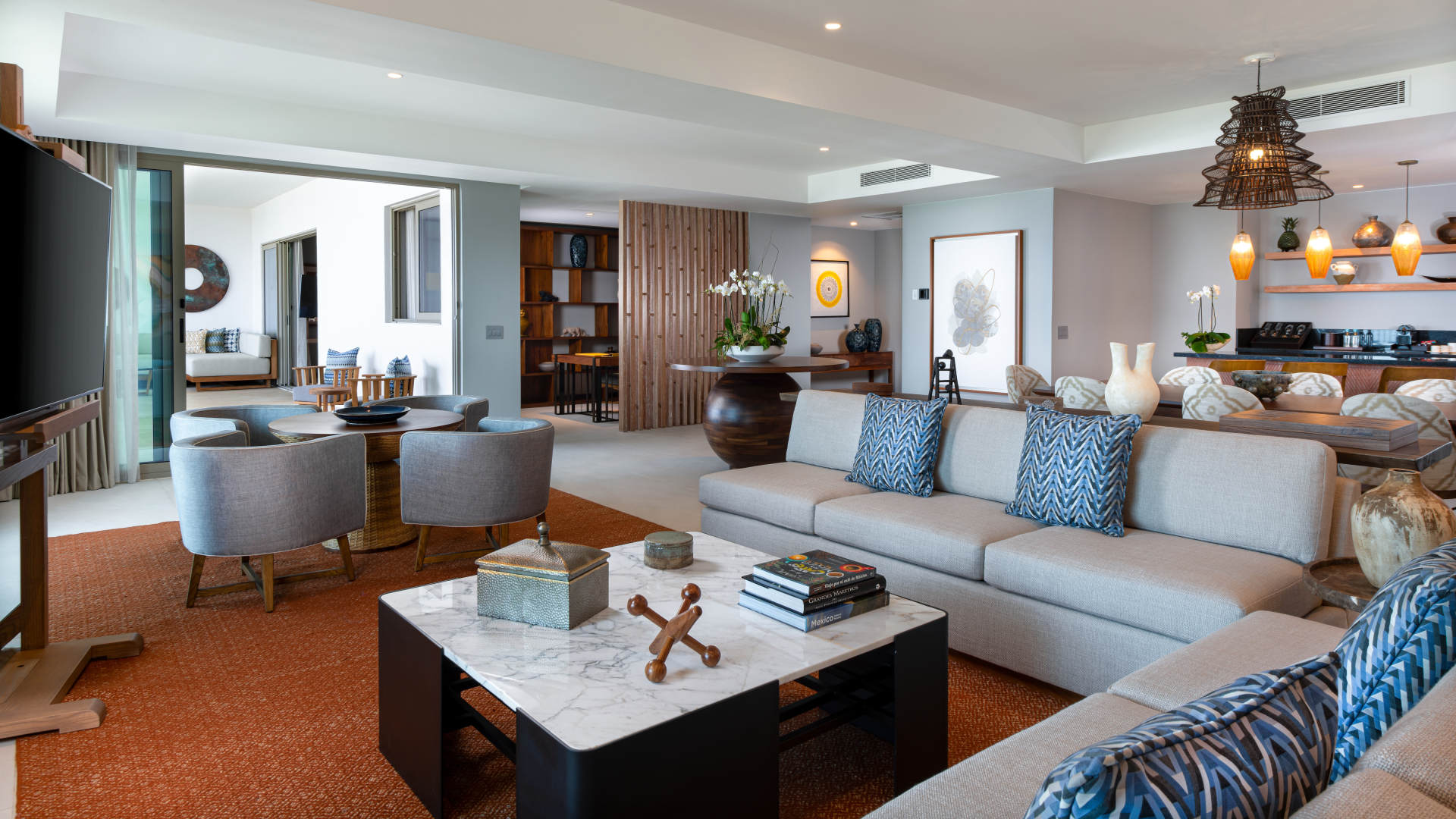 Spacious and modern living room with neutral-colored sofas, blue patterned pillows, a marble coffee table with decorative items, and a large flat-screen TV. In the background, there is a dining area with a wooden table and chairs, decorative vases, artwork on the walls, wooden shelves with ornaments, and a view into a bright sunroom with outdoor seating.