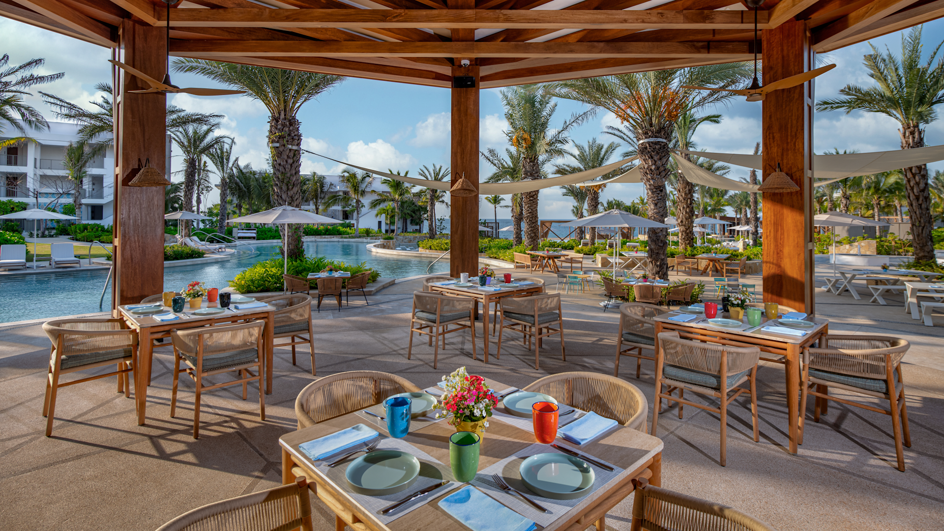 Outdoor poolside dining area with tables set with plates, cups, napkins, and flowers, surrounded by palm trees and lounge chairs, under a wooden canopy.