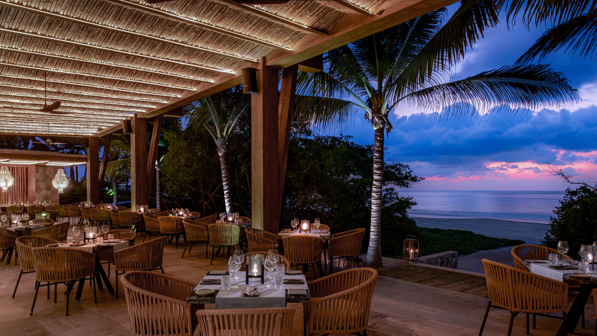 Outdoor restaurant with tables set for dining, surrounded by palm trees, overlooking the ocean at sunset, with blue and pink sky.