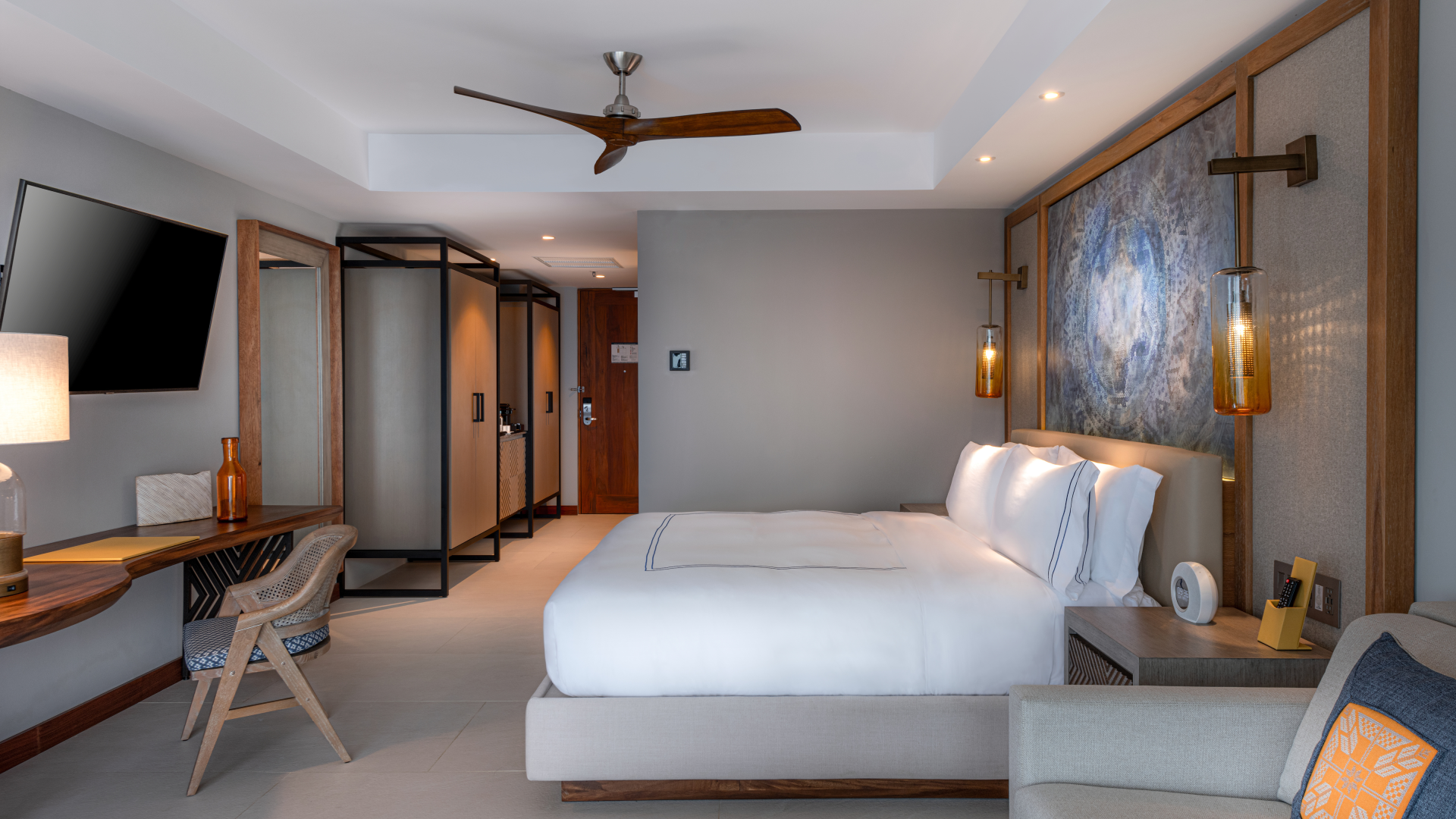 Modern hotel room with a king-sized bed, a large flat-screen TV on the wall, a wooden desk with a chair, a decorative wall panel behind the bed, bedside lamps, and a sitting area with pillows.