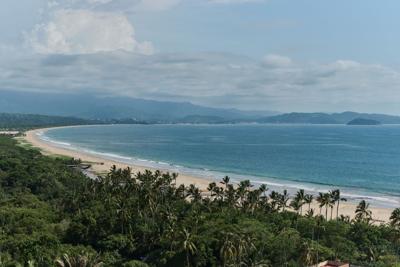 Scenic view of a tropical beach with palm trees, sandy shoreline, and ocean under a partly cloudy sky, with mountains in the background.