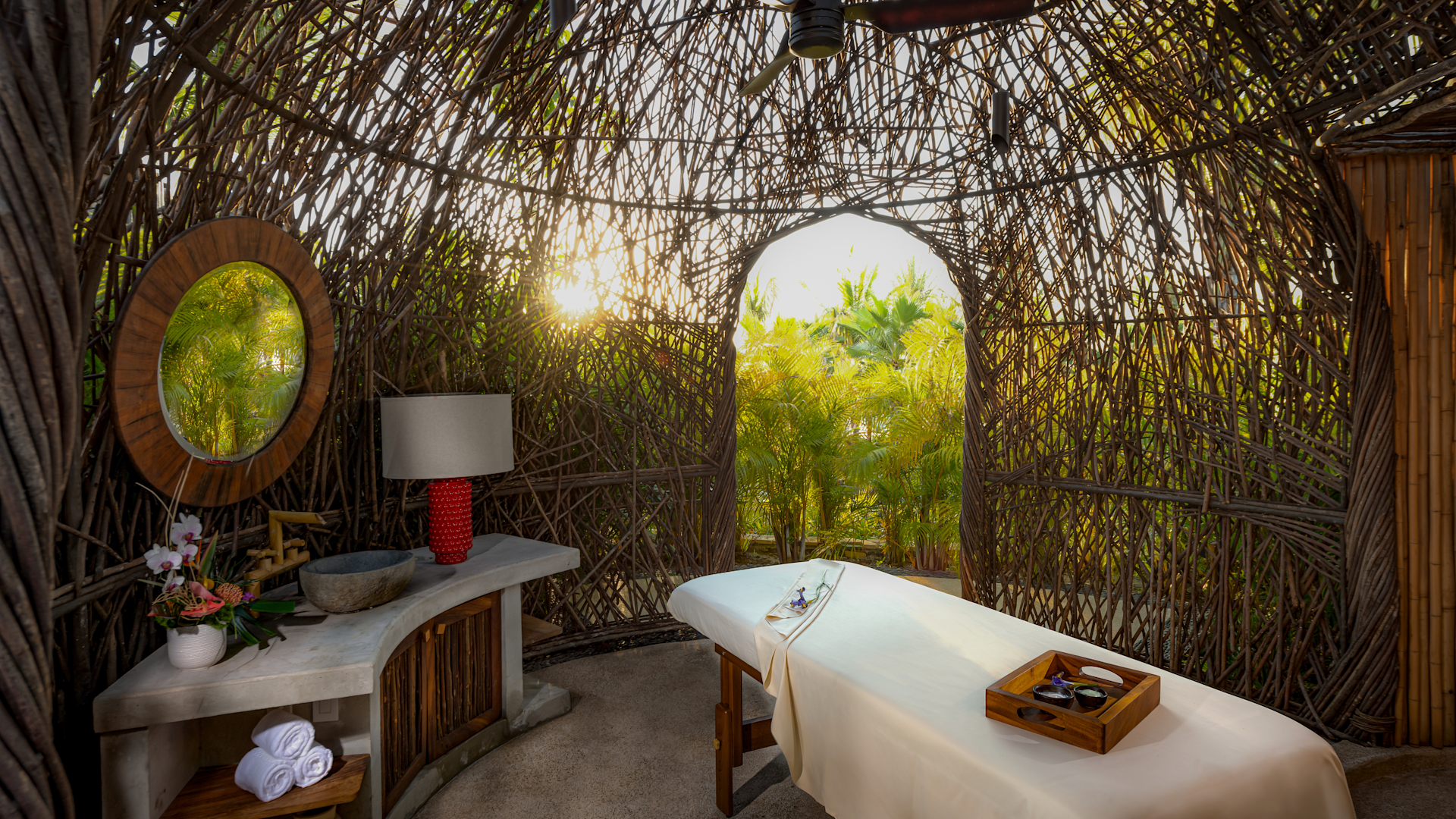 A tropical spa treatment room with a massage table covered in a white sheet, surrounded by a woven wicker wall with an opening showing lush green foliage and sunlight outside. There is a small table with a flower arrangement, a lamp, and a stone bowl inside the room.