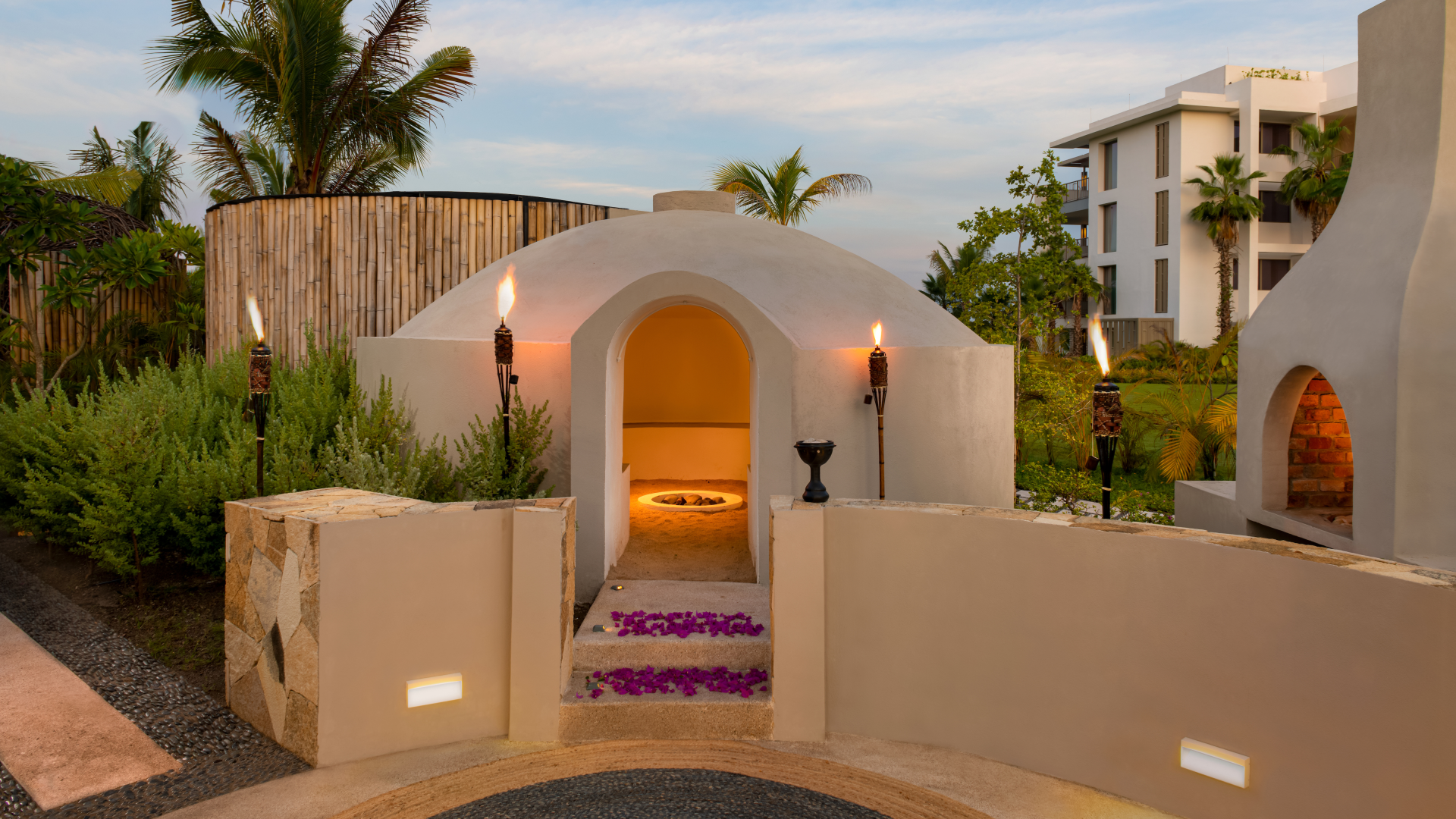A white, dome-shaped outdoor fireplace area at sunset, decorated with tiki torches, purple flower petals on steps, and surrounded by tropical plants and modern buildings.
