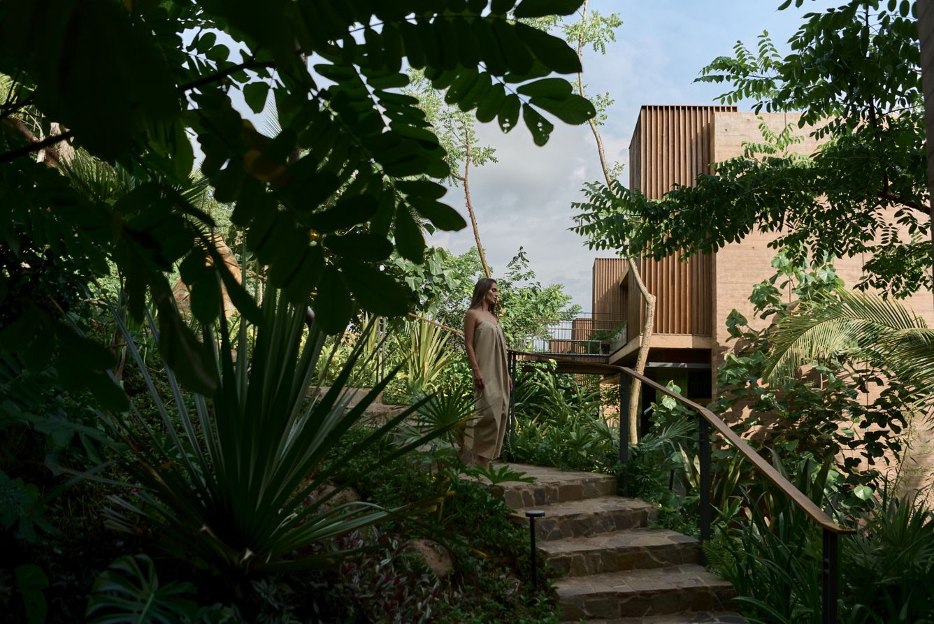 A woman in a beige dress standing on stone steps surrounded by lush green tropical plants and trees, with a modern building in the background under a partly cloudy sky.
