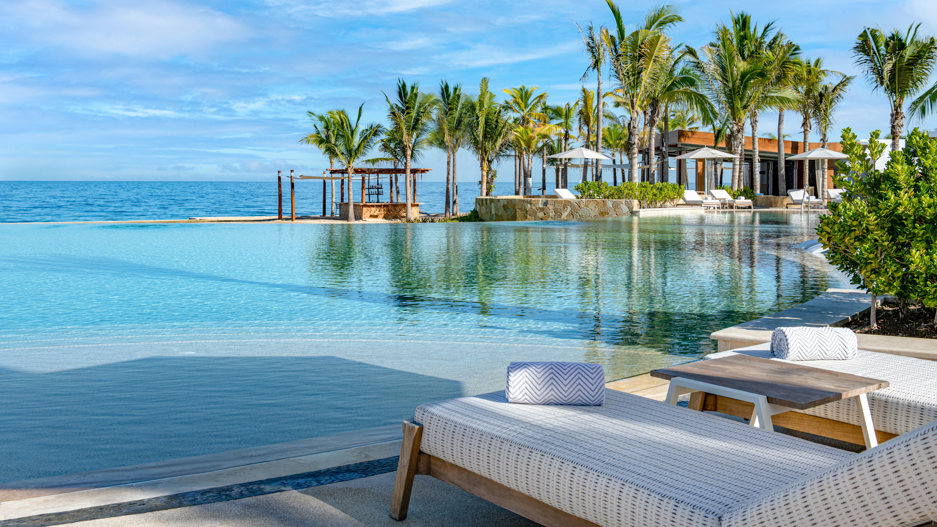 Luxury beachfront resort with swimming pool, palm trees, lounge chairs, umbrellas, and ocean view.