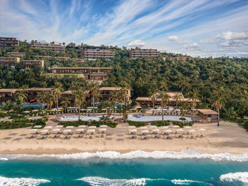 Luxury beachfront resort with multiple buildings on a hillside, palm trees, a pool area, and a sandy beach with lounge chairs and umbrellas, overlooking the ocean under a partly cloudy sky.