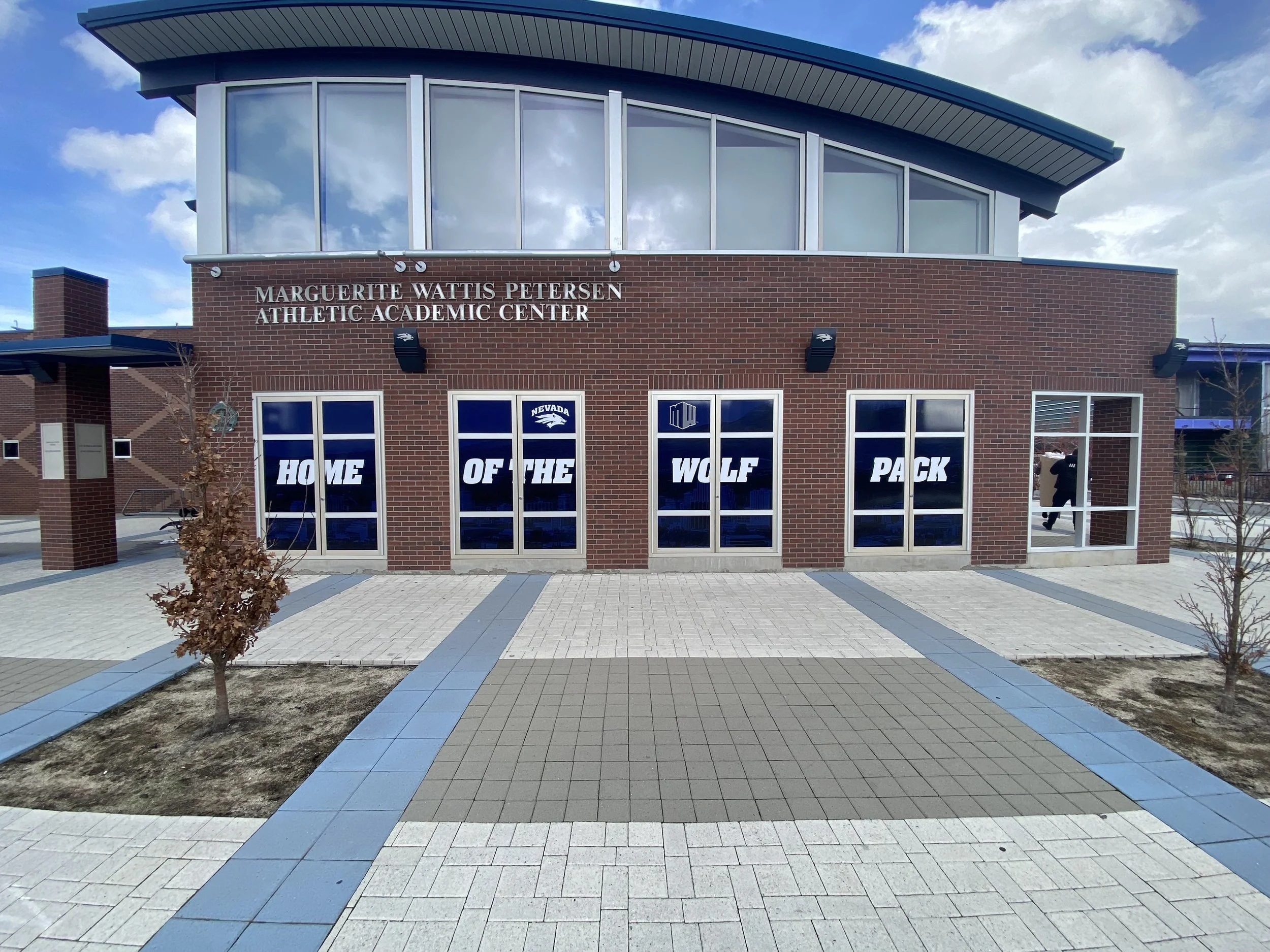 Front view of the Marguerite Watts Petersen Athletic Academic Center with a brick exterior, large windows with a blue banner reading 'Home of the Wolf Pack', and a sidewalk with trees.