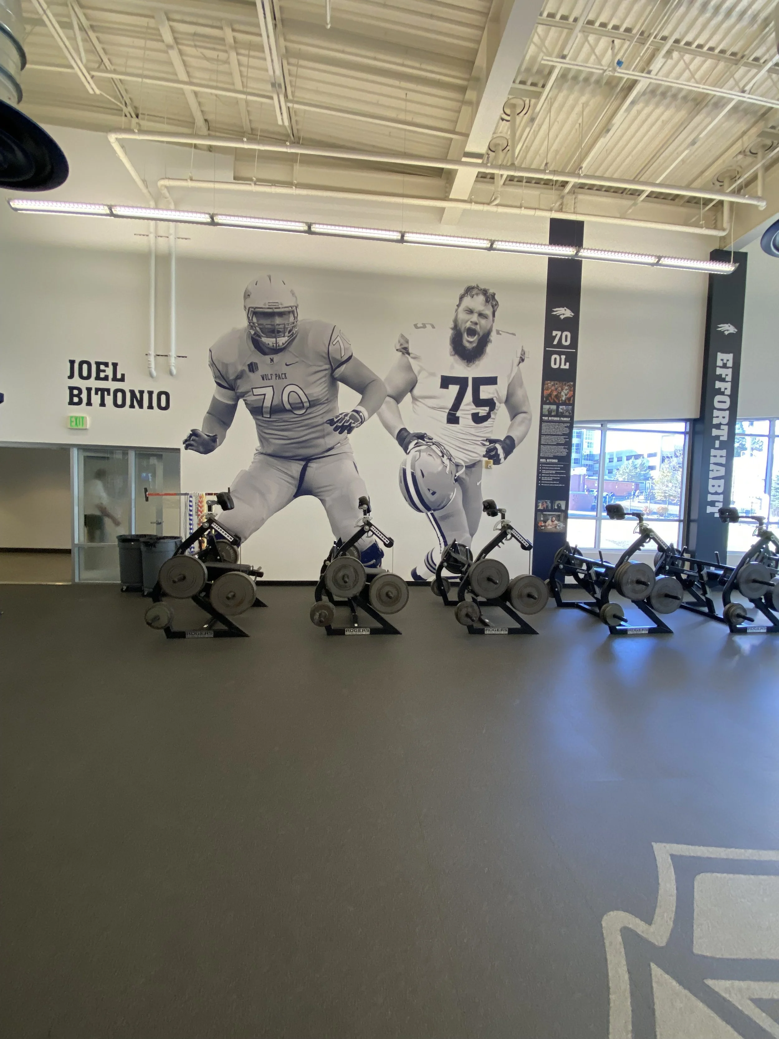 Large mural of two football players in black and white, one wearing a helmet and the other with a beard, both in action poses, behind a row of exercise bikes in a gym.