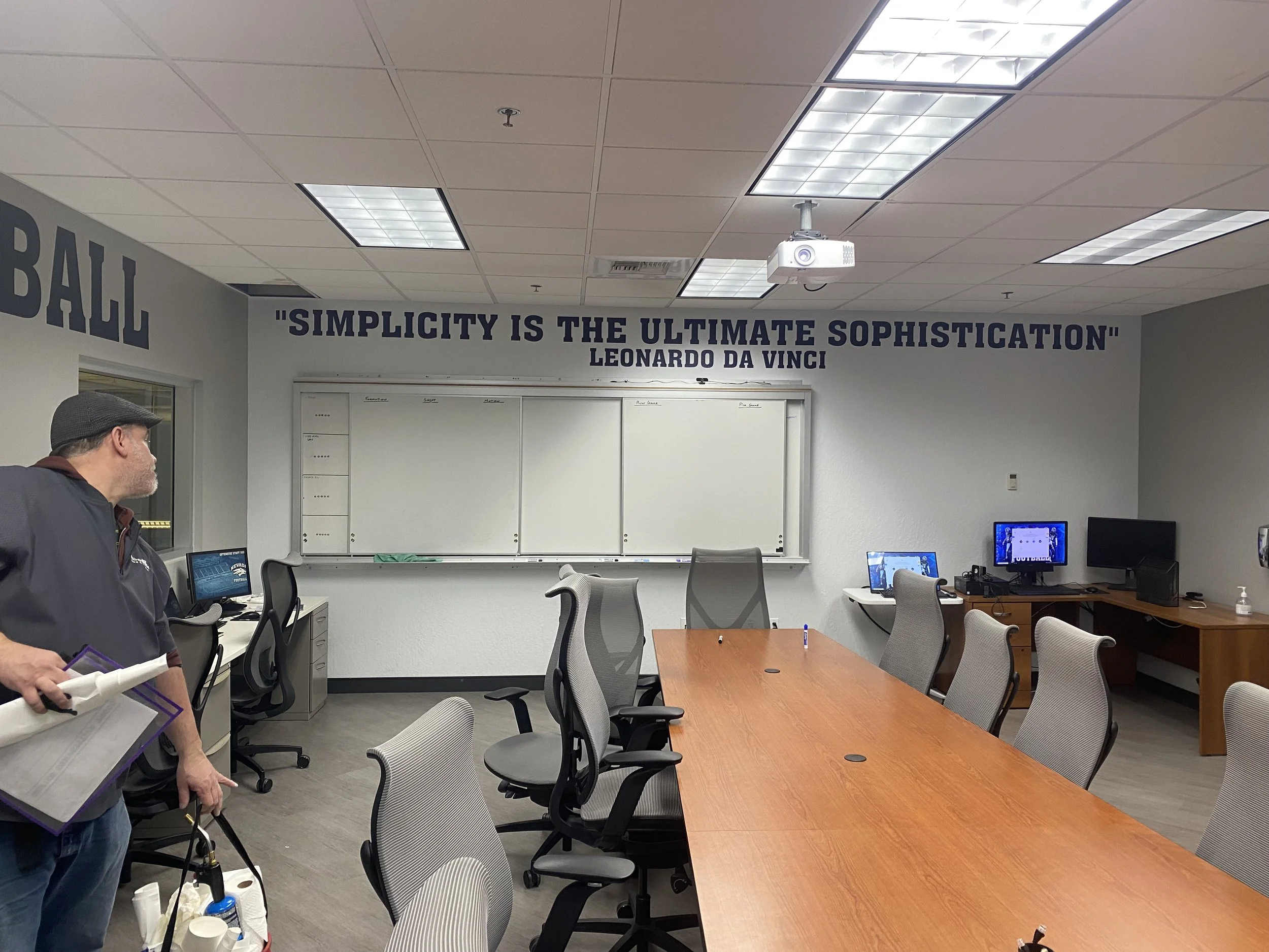 Empty conference room with a quote on the wall saying 'Simplicity is the ultimate sophistication' by Leonardo da Vinci, whiteboard, several chairs around a wooden table, computers on desks, and a person holding cleaning supplies.