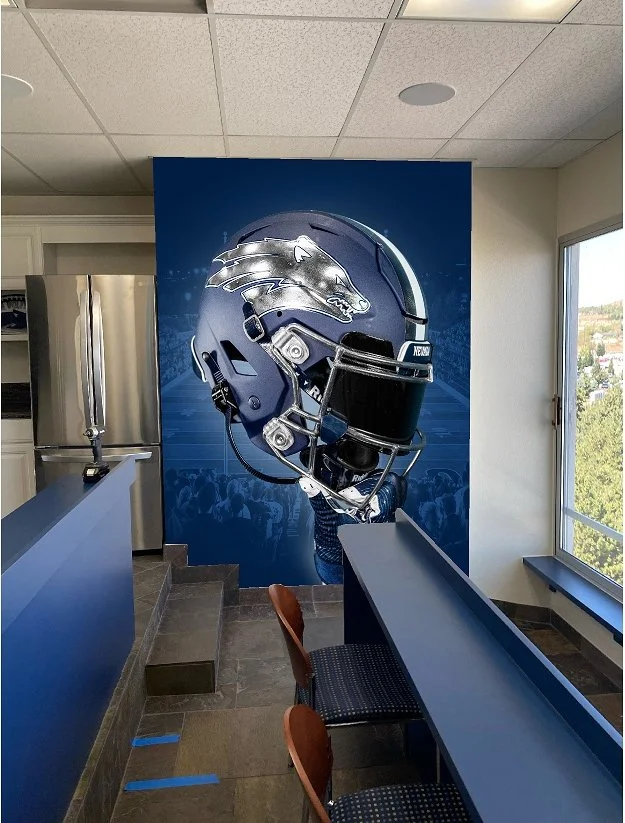 Interior of a sports bar or restaurant with a large Denver Broncos helmet mural on the wall, a counter with bar stools, a window showing trees outside, and a refrigerator in the corner.