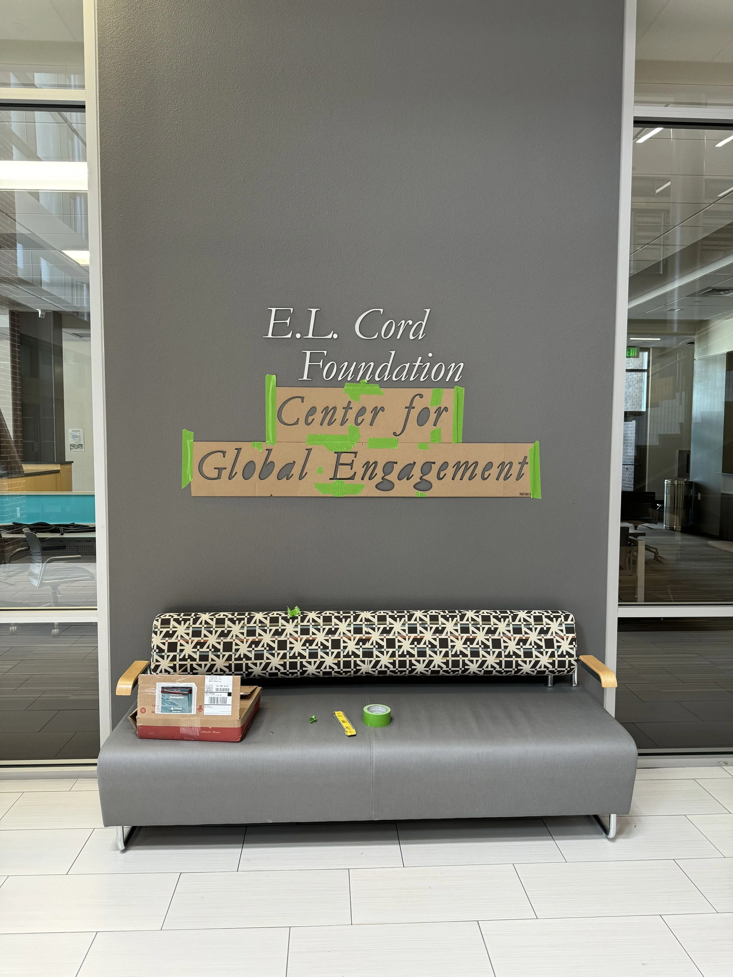 Interior view of a modern office or lobby area with a gray wall displaying a sign for the E.L. Cord Foundation Center for Global Engagement, handwritten on taped cardboard. A gray couch with a patterned bolster pillow in front of the wall, with a box