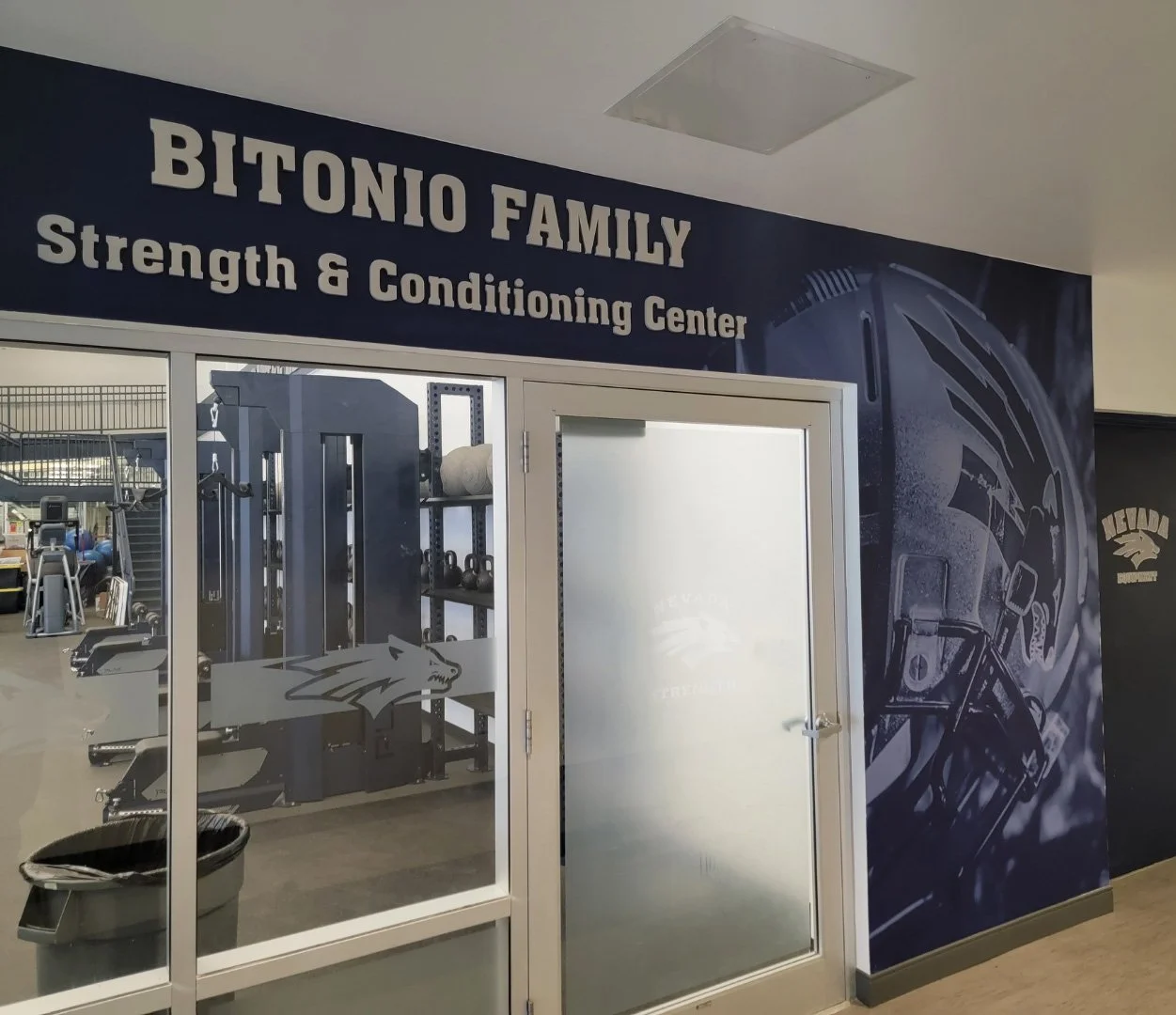 Entrance to the Bitonio Family Strength & Conditioning Center with gym equipment visible inside and a wall graphic featuring a football helmet and other sports equipment.