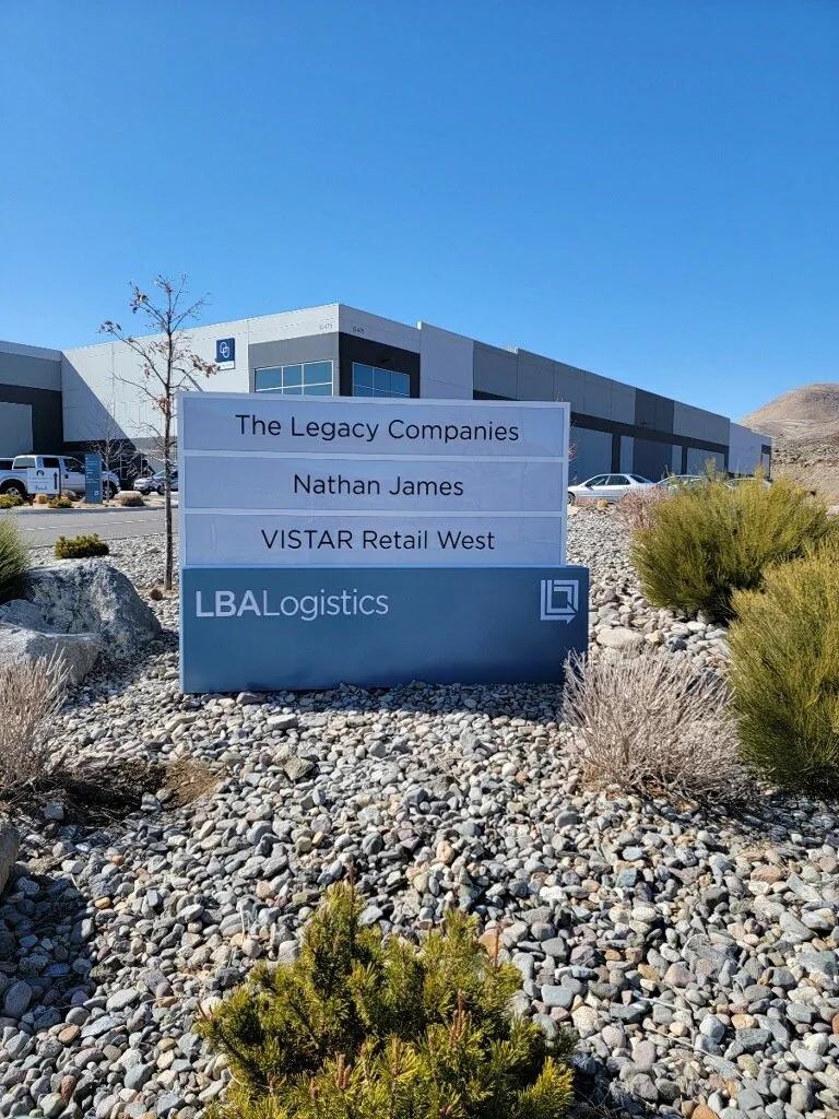 Outdoor sign for LBA Logistics with businesses listed: The Legacy Companies, Nathan James, VISTAR Retail West, in front of a commercial building, with a parking lot and rocky landscaping, under a clear blue sky.