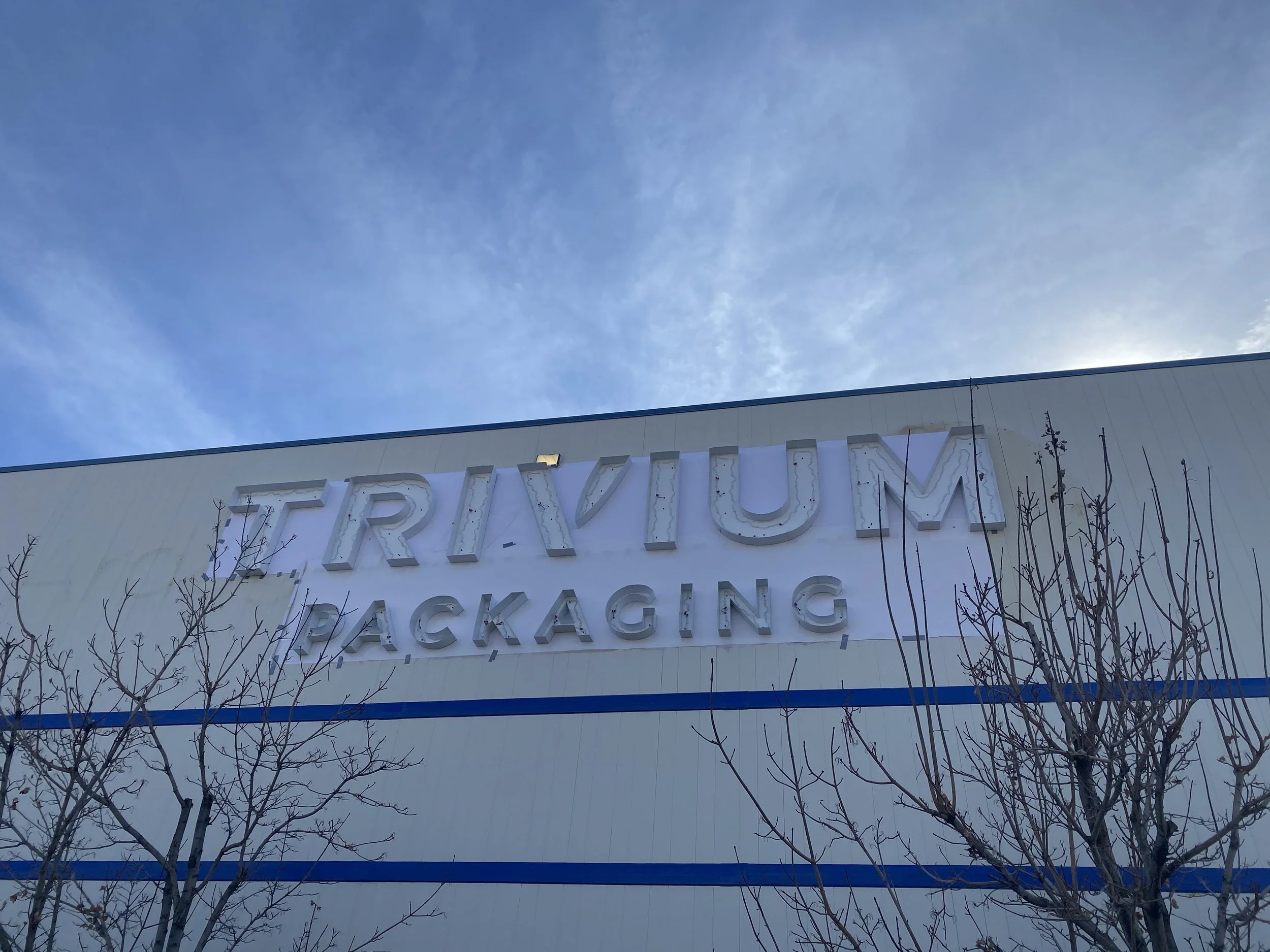 Exterior of a large building with a sign that reads 'TRIUM PACKAGING' against a blue sky with some clouds and bare trees in the foreground.