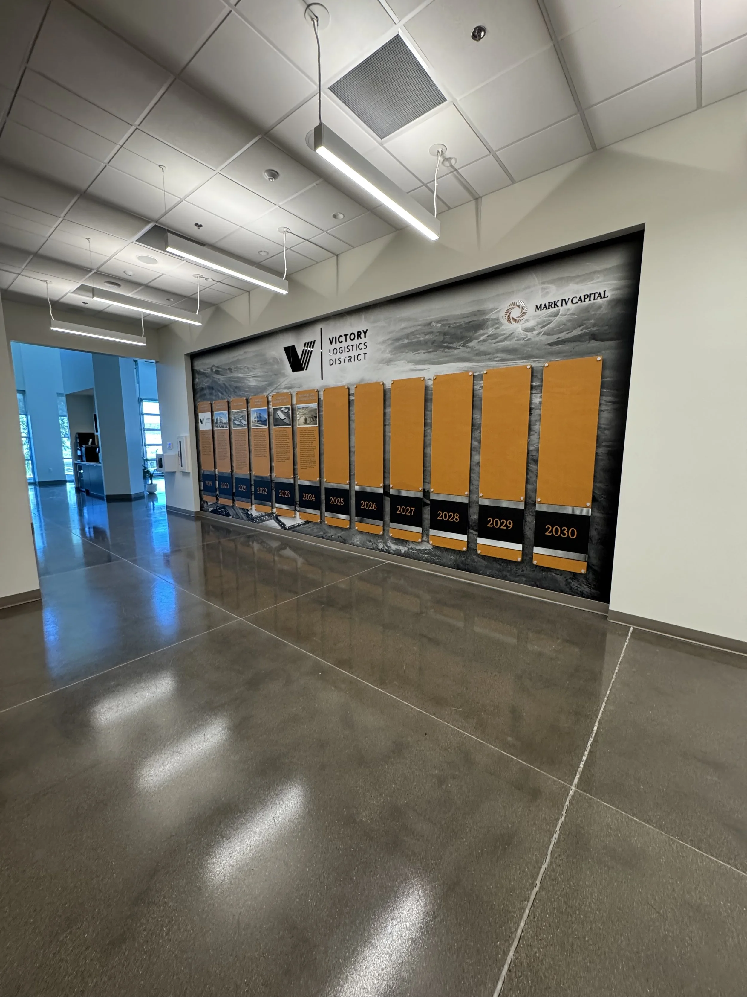Office lobby with a timeline display on the wall labeled 'Victory Logistics District' and 'Market Capital,' showing years 2000 to 2030, with orange panels and descriptive text.
