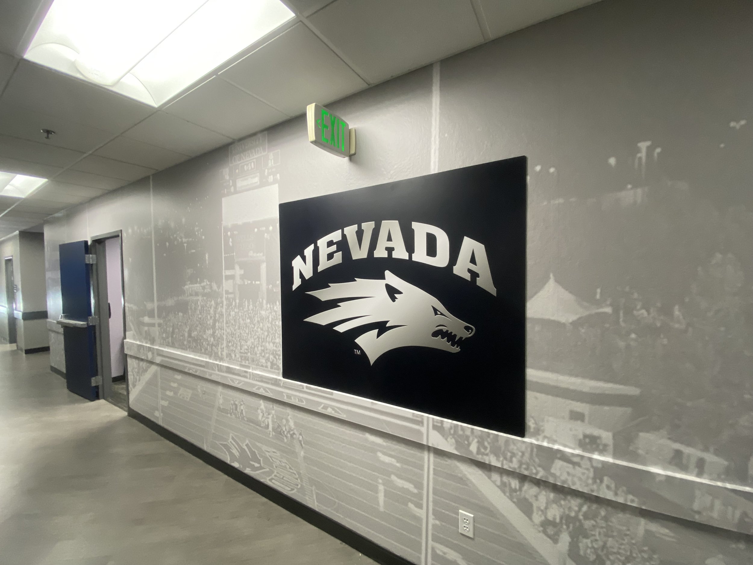Indoor hallway with a large Nevada logo featuring a wolf's head on the wall and an emergency exit sign above.