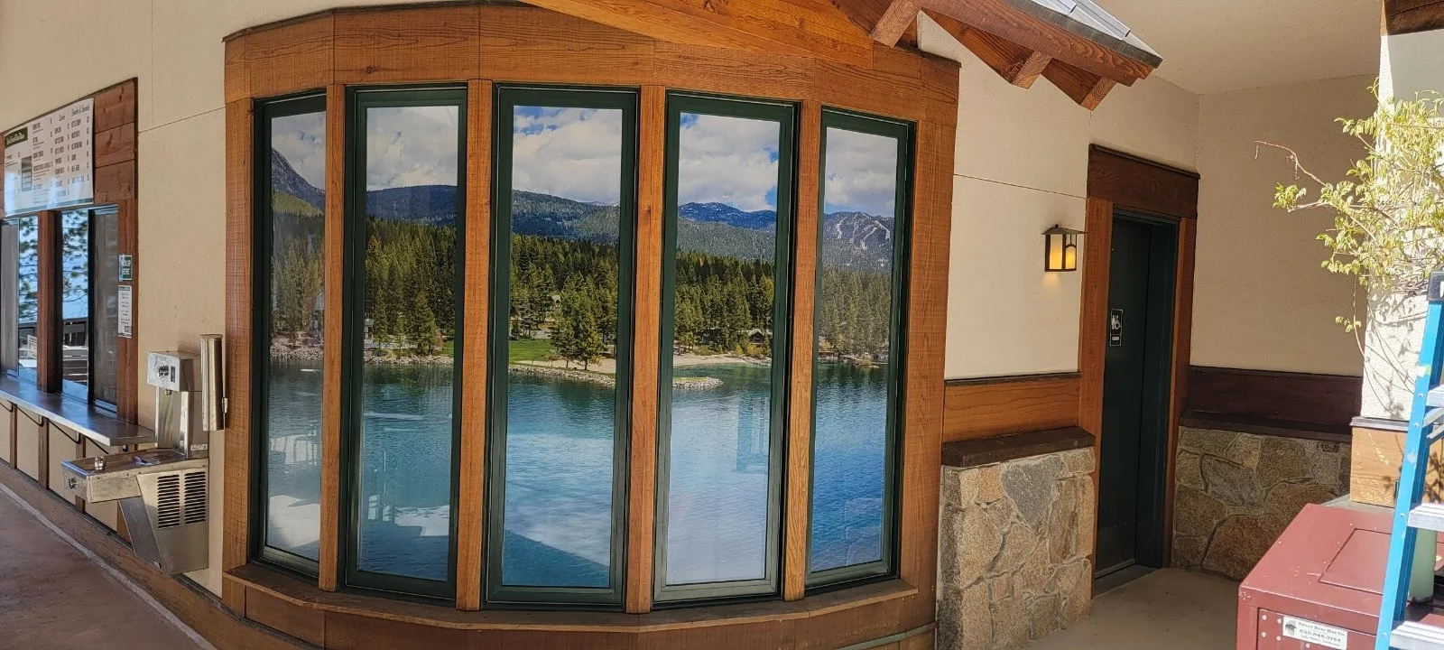 Indoor view of a building with large glass window reflecting a lake, mountains, and sky outside, wood and stone accents, and a water fountain on the wall.