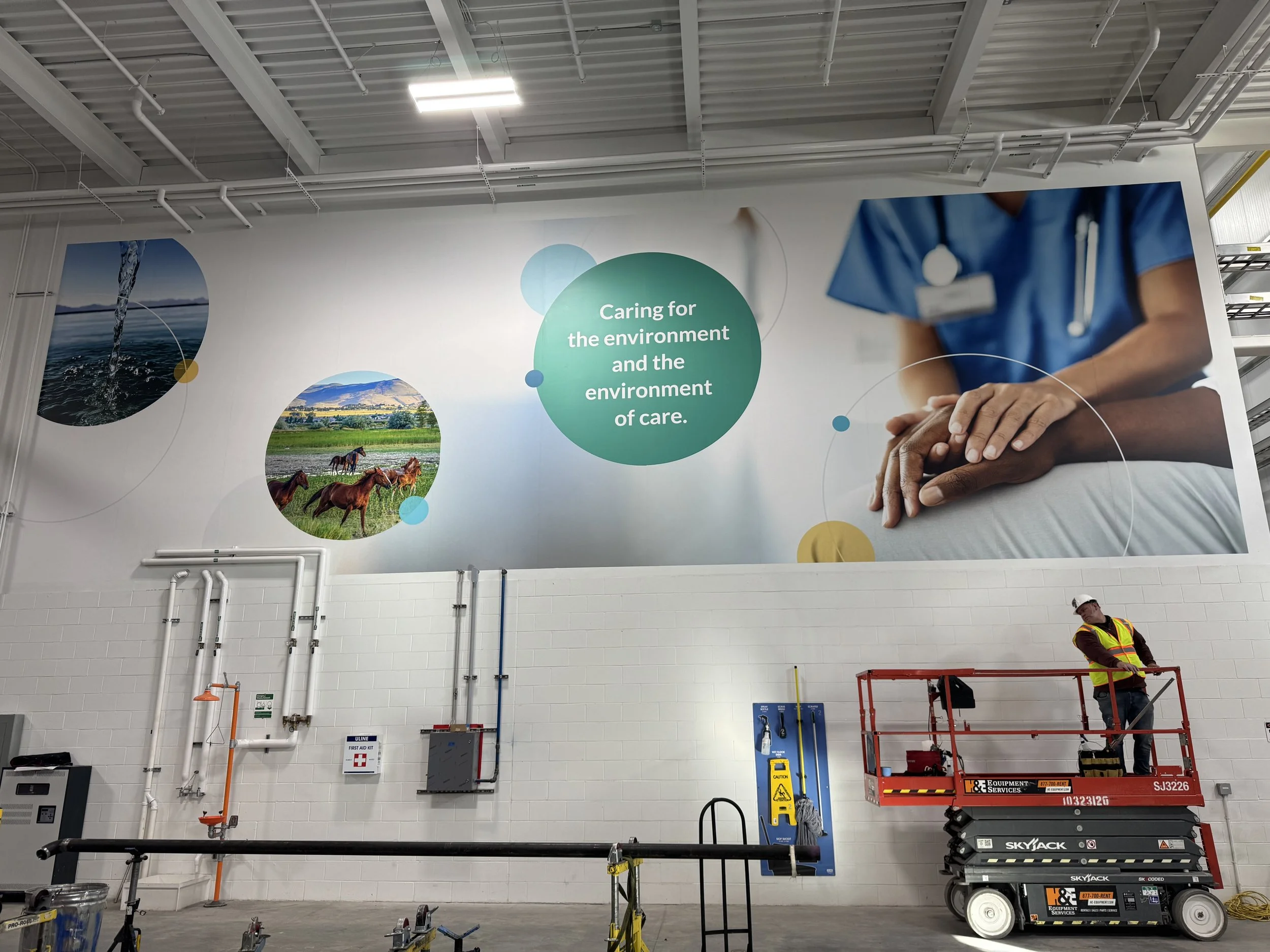 Large interior warehouse with a high ceiling and a prominent wall mural showing nature and healthcare themes. A worker in a yellow vest and white helmet is standing on a red scissor lift near wall-mounted equipment.
