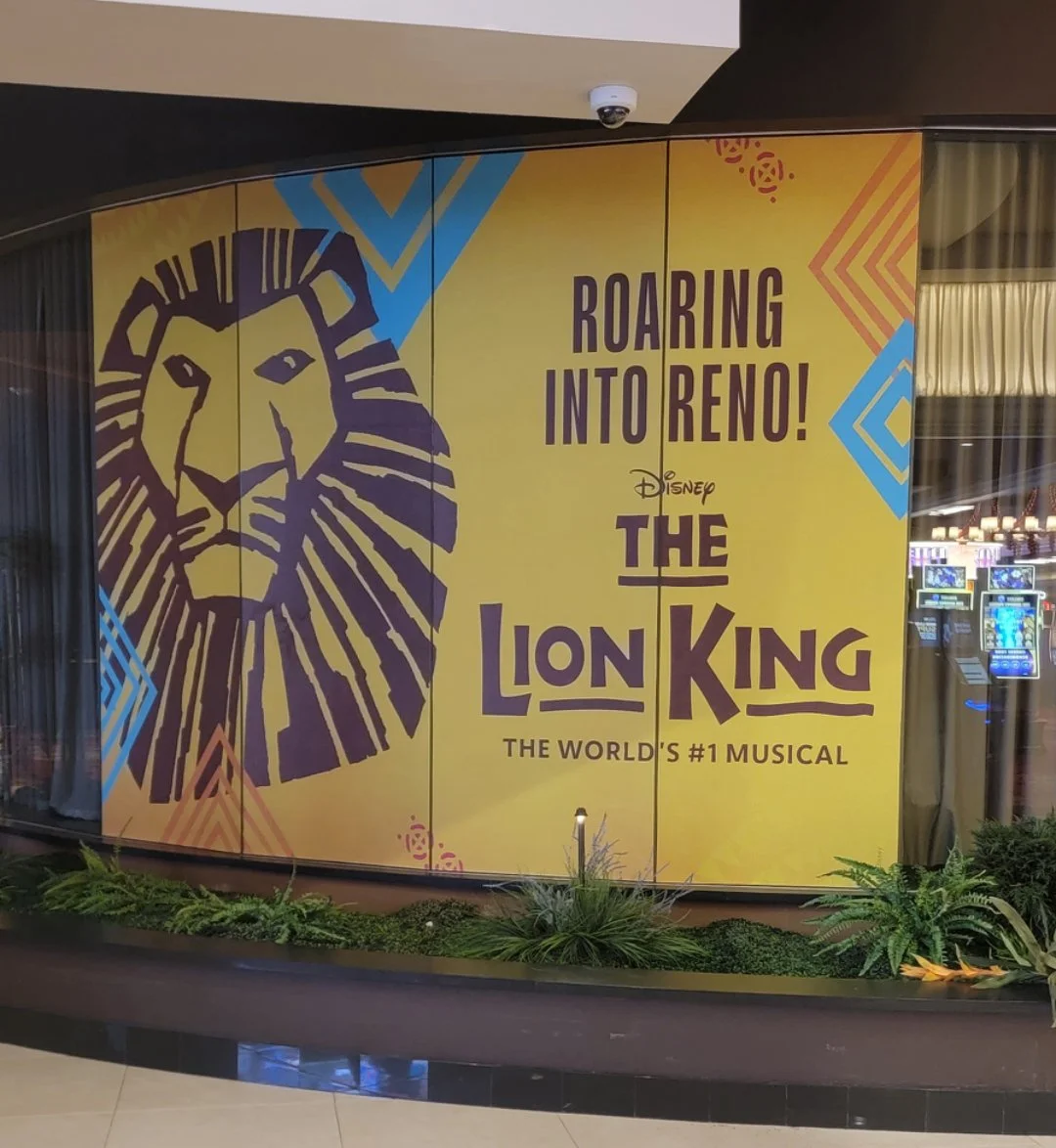 Poster advertising Disney's The Lion King musical with a large stylized lion's face on the left and the text 'Roaring Into Reno! The Lion King The World's #1 Musical' on a yellow background.