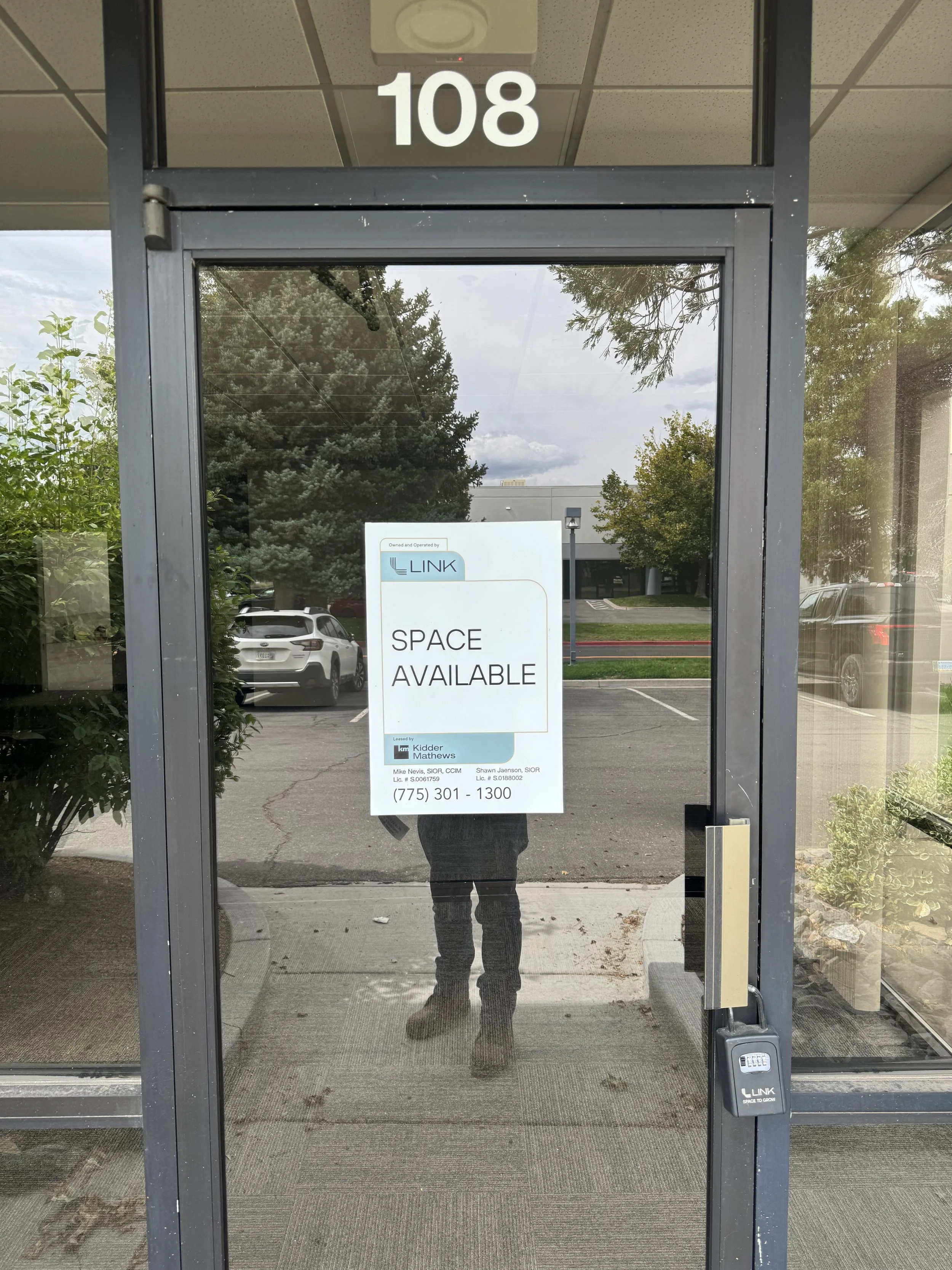 Glass door with a sign that reads 'Space Available' and the number 108 above it. Reflection of the person taking the photo, parking lot, trees, and sky outside can be seen in the glass.