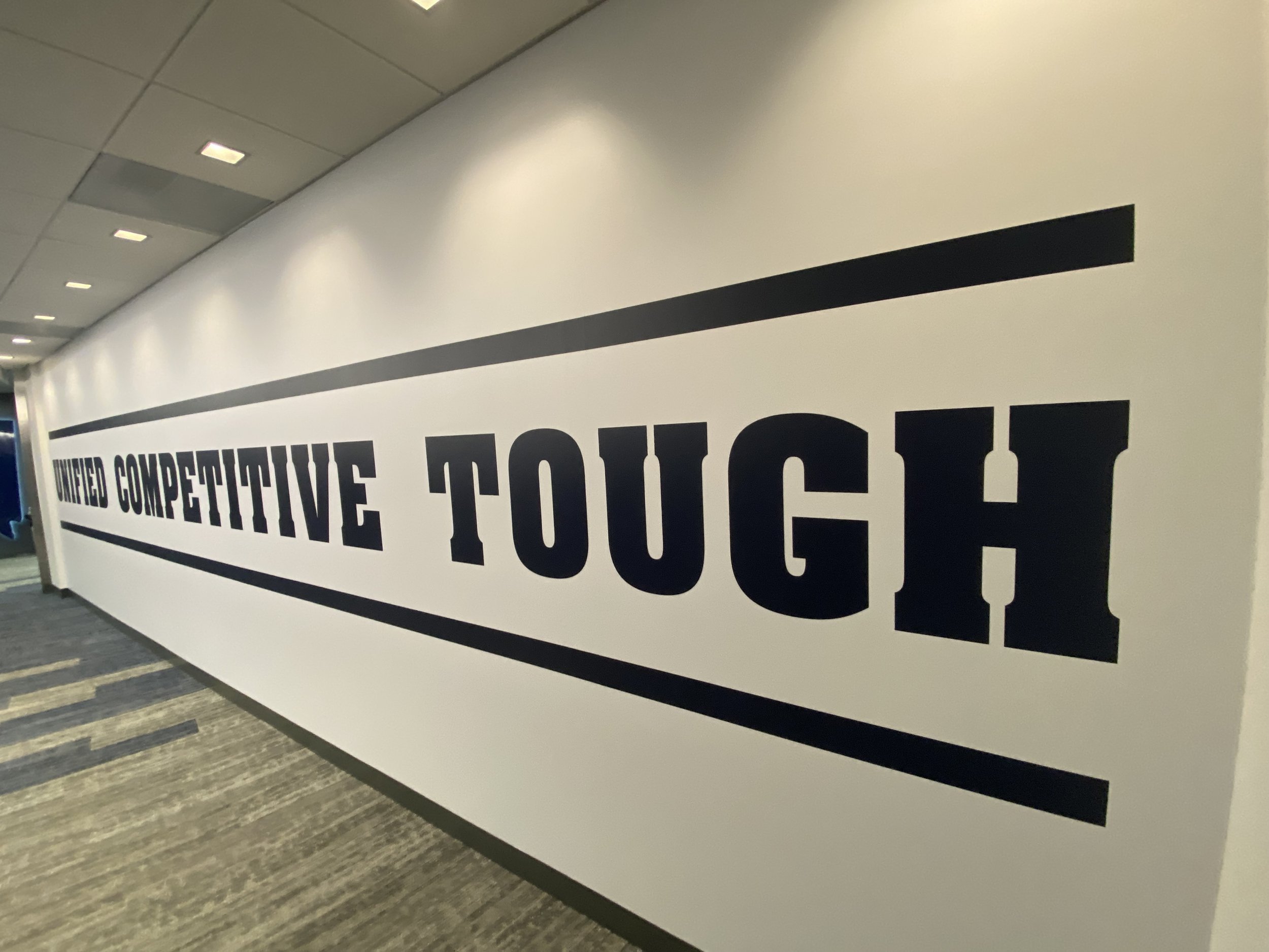 A white wall with black text that says 'United Competitive Touch' in a hallway with a gray patterned carpet and a ceiling with recessed lighting.