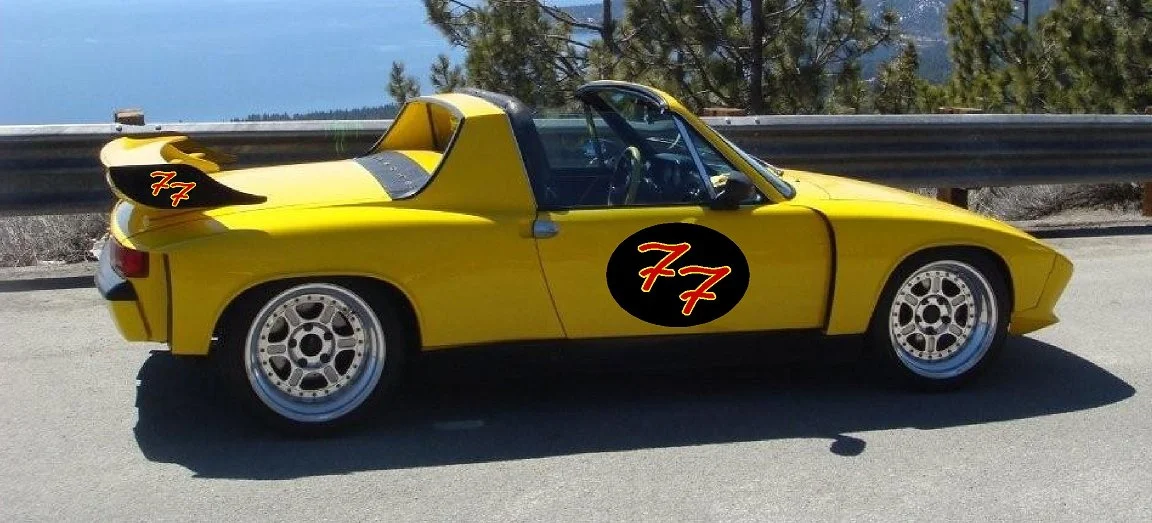 A yellow sports car with custom black and red decals that say '77', parked on the side of a mountain road with trees and mountains in the background.
