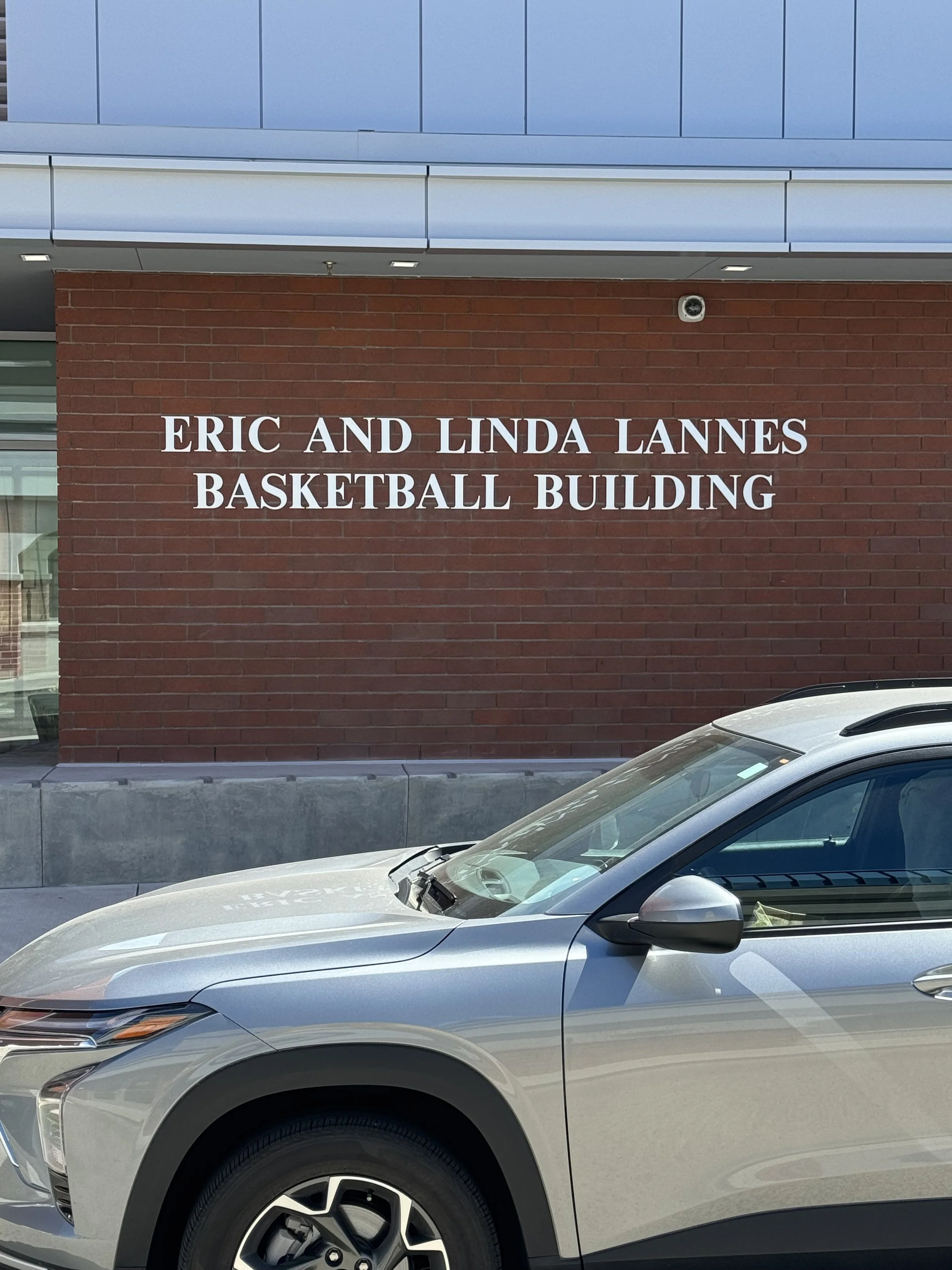 Brick wall with sign reading 'Eric and Linda Lannes Basketball Building'. Part of a silver car is visible in the foreground.