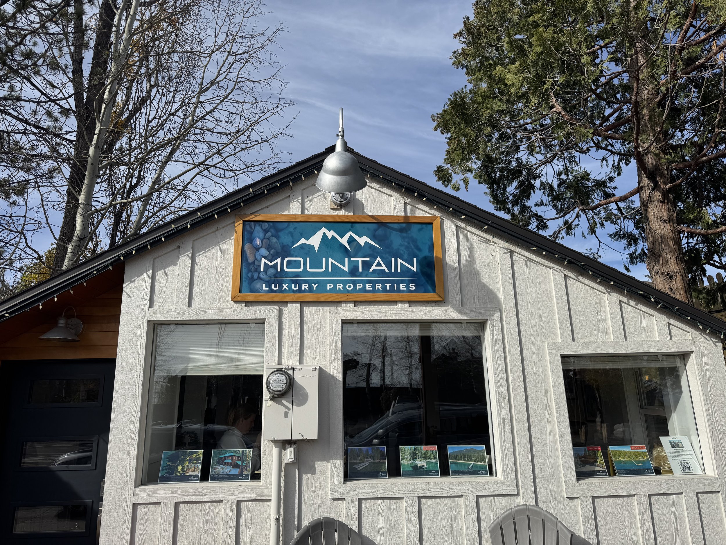Front view of a building with a sign that says 'Mountain Luxury Properties,' surrounded by trees, with a clear sky overhead.