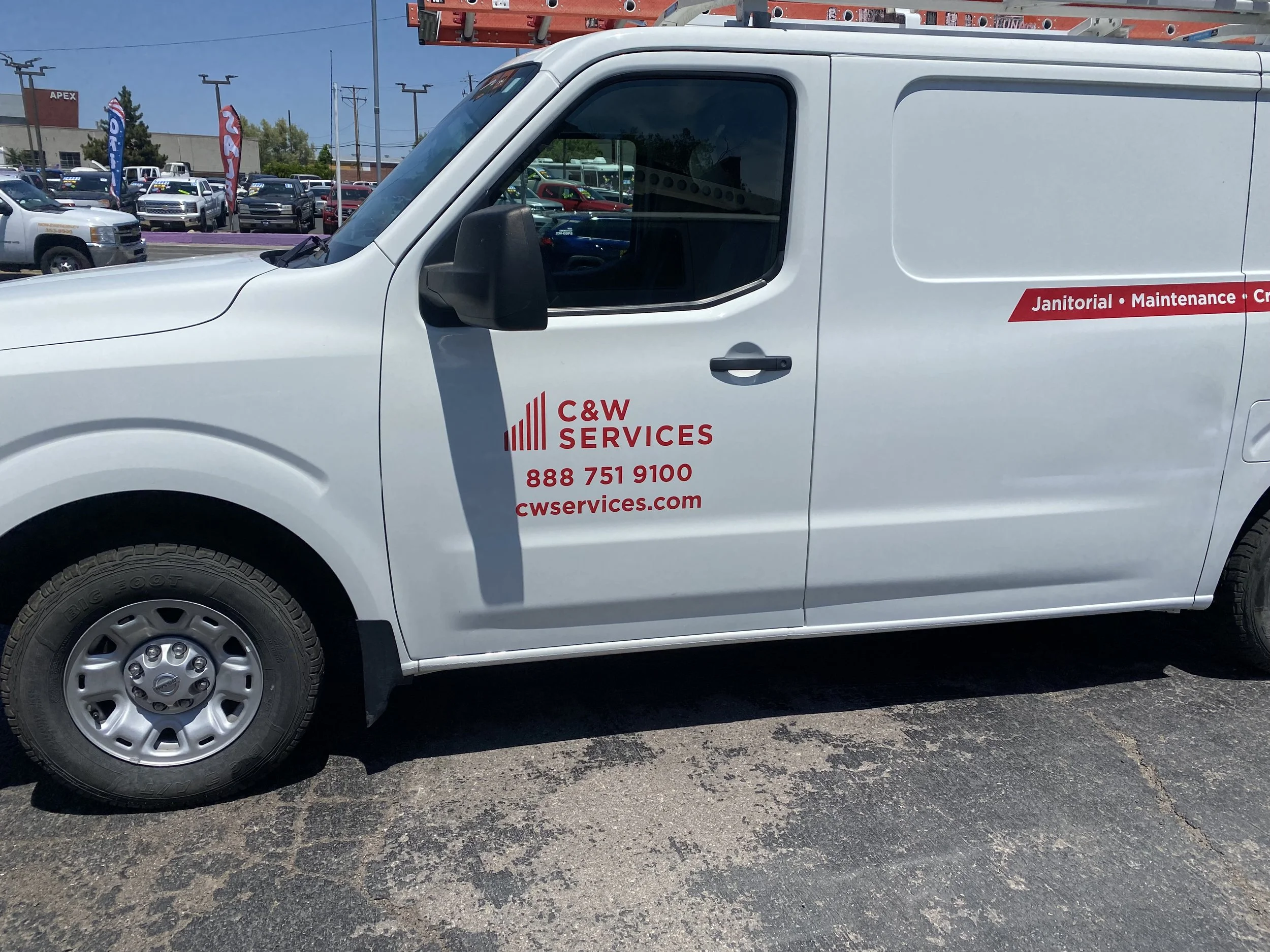 White service van with red lettering and branding for C&W Services, parked in a lot with other vehicles and a building in the background.