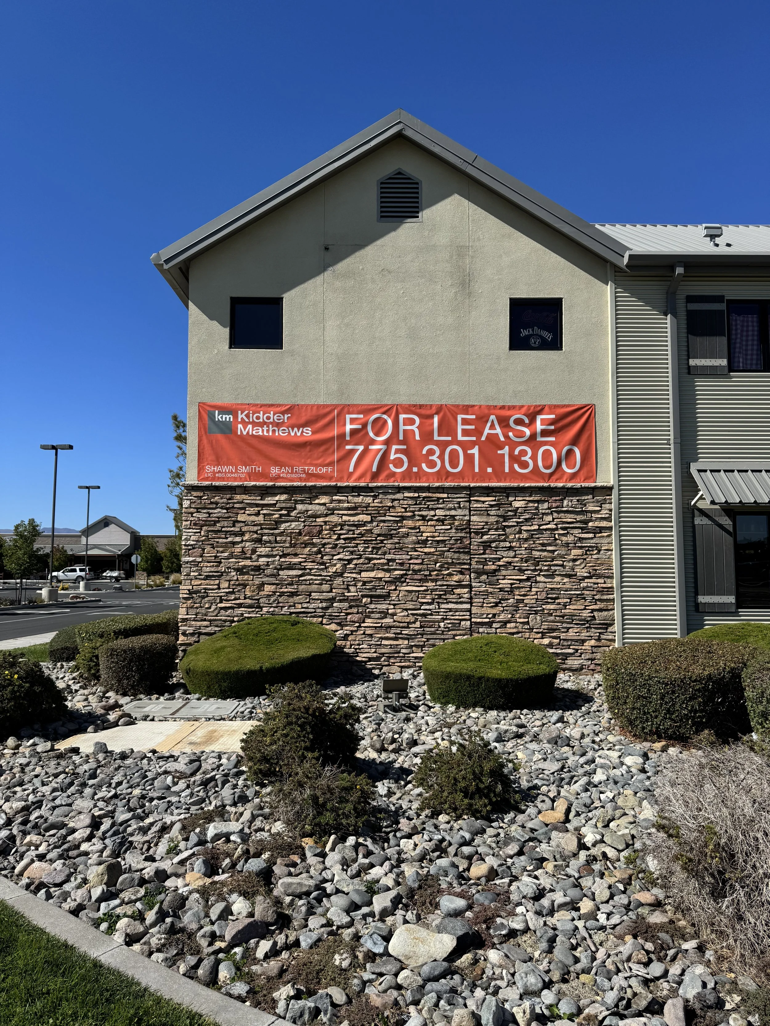 A two-story building with a stone foundation and beige upper walls, featuring a large red banner advertising the property for lease with contact information. The building has a small shrub and rock landscaping in front, and the sky is clear and blue.