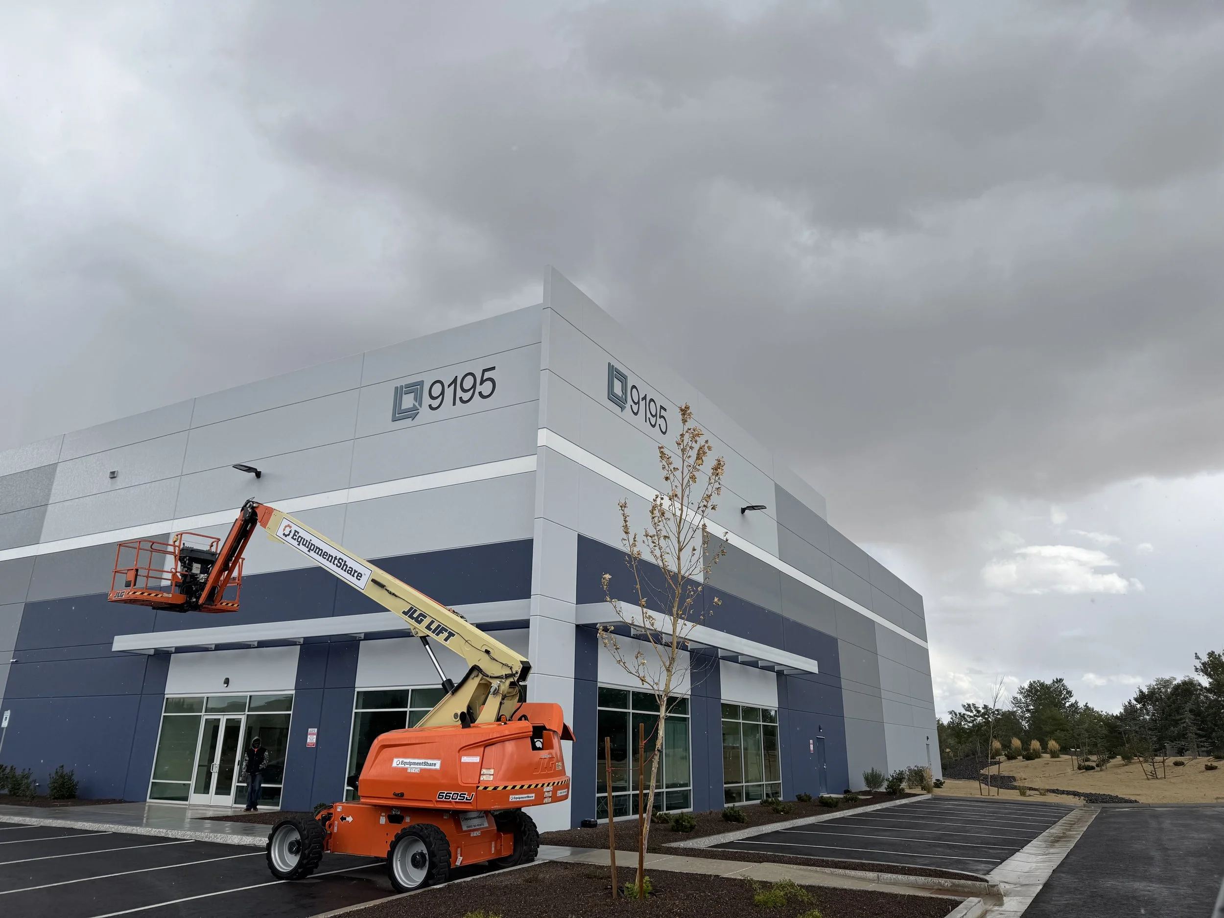 A modern commercial building under dark, cloudy skies with a cherry picker lift in the parking lot, and a young tree planted nearby. There is a lift for the SilverBlu to make a sign install.