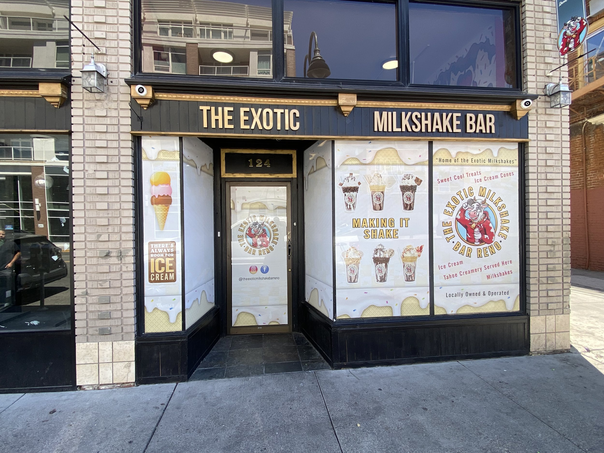 Front view of an ice cream shop called 'The Exotic Milkshake Bar' with signage advertising ice cream and milkshakes. The storefront has large windows with promotional graphics of milkshakes and ice cream cones. The entrance is centered, with the shop