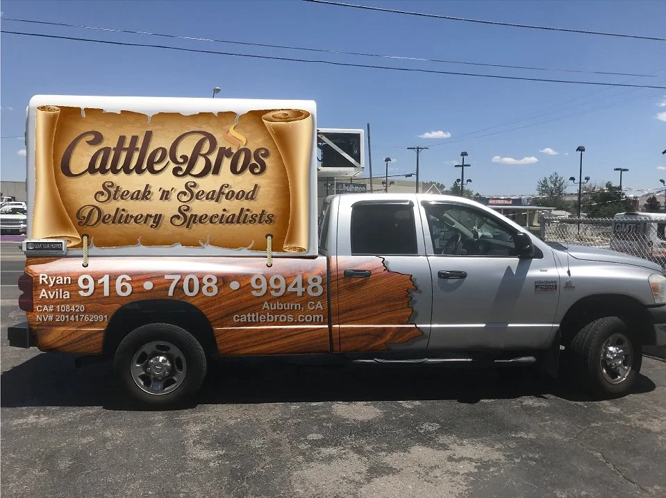 A pickup truck with an advertisement for Cattle Bros steak and seafood delivery service. The truck's side features a large sign with a rolled paper design and text, as well as a graphic of the state of California with a wood grain pattern.