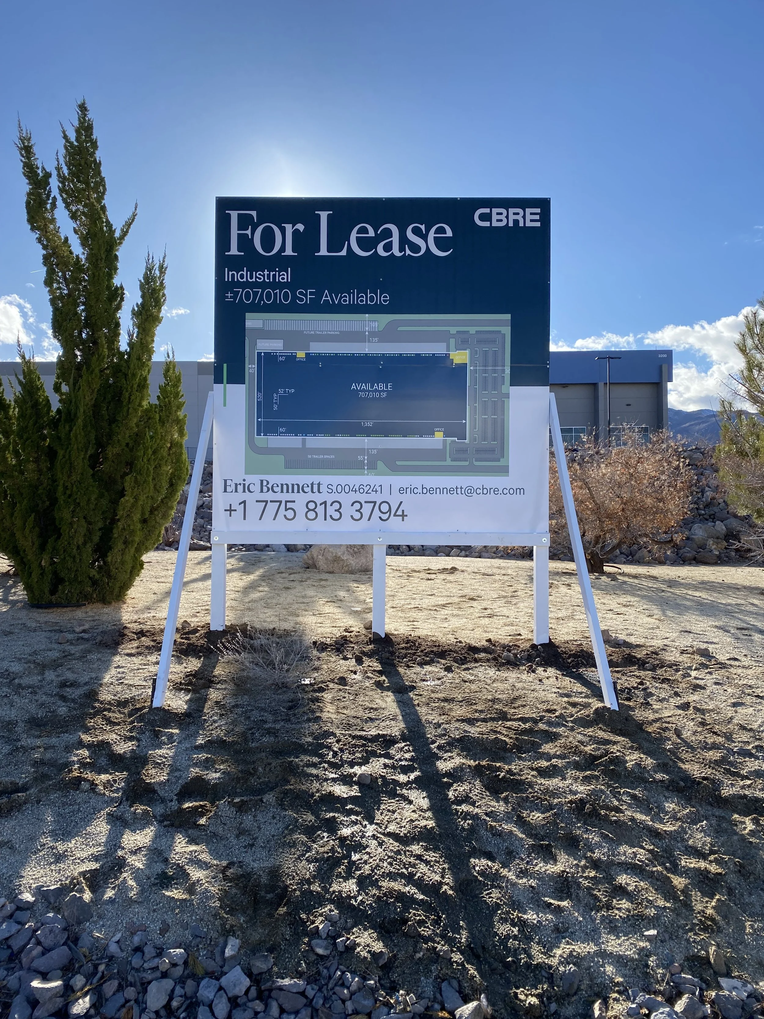A large outdoor signboard indicating a commercial property for lease, showing a detailed site plan and contact information for Eric Bennett, with a clear sky and mountains in the background.