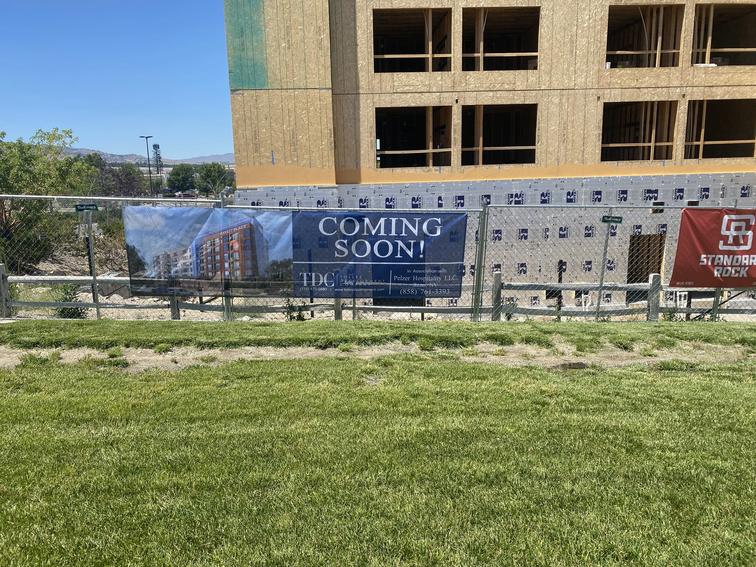 Construction site with a building under construction and a sign that reads "Coming Soon!" with an image of the future building. There is a grassy foreground and a chain-link fence around the site.