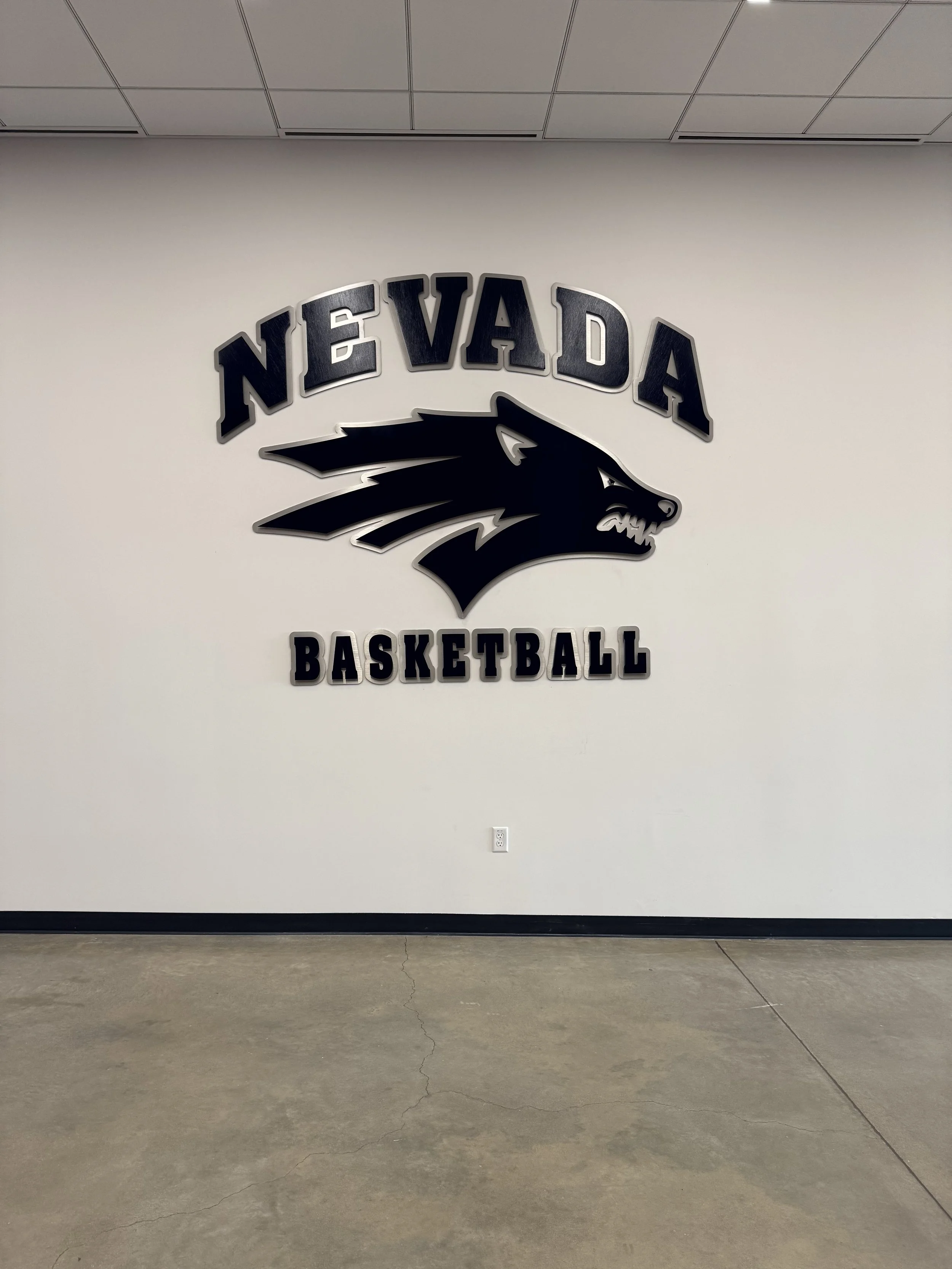 Wall with a large black and silver Nevada basketball team logo featuring a wolf's head and the words 'NEVADA BASKETBALL' on a white wall.